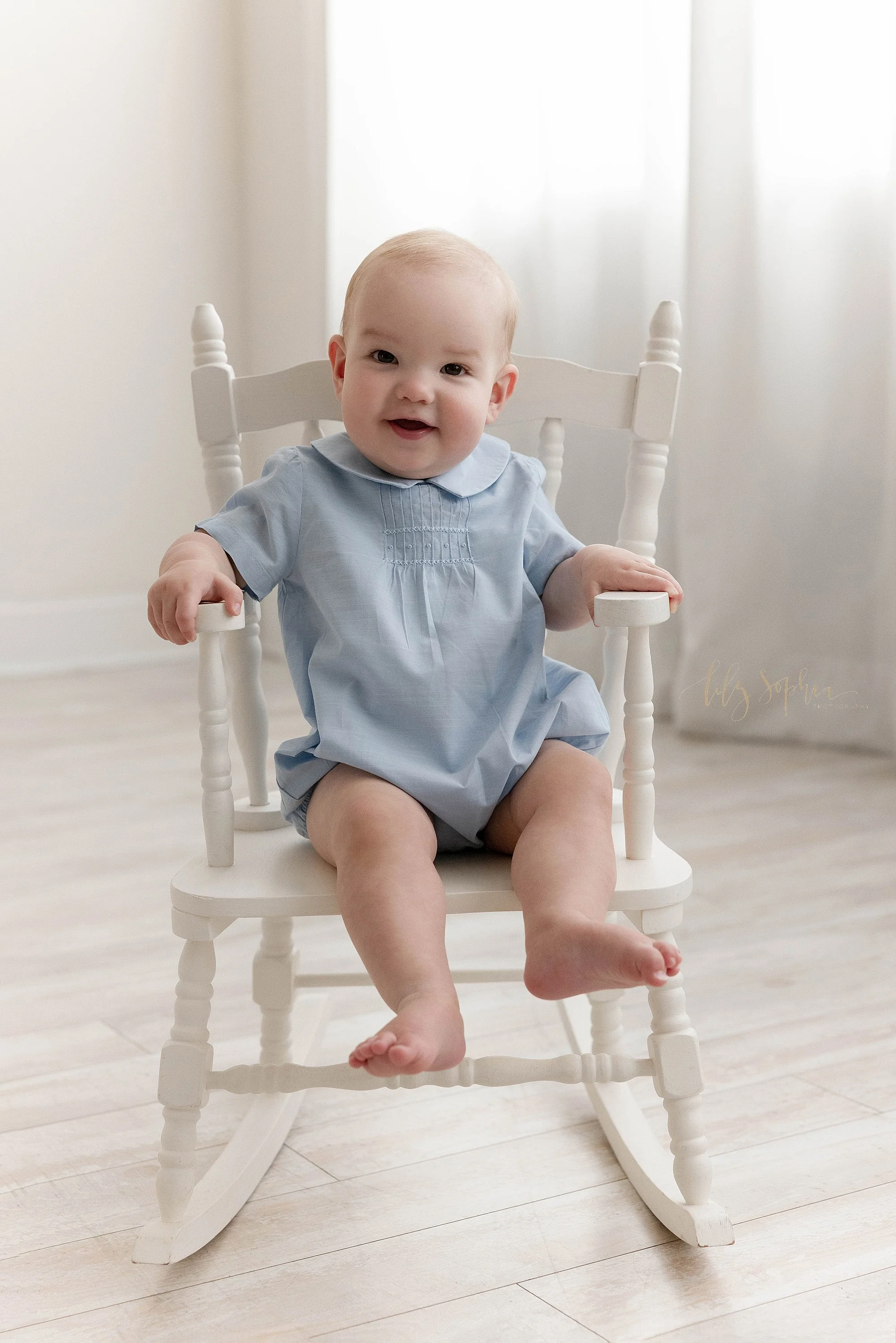  A one year old baby boy sits in a rocking chair in front of window streaming natural light for a first birthday photography session in a studio near Kirkwood in Atlanta. 