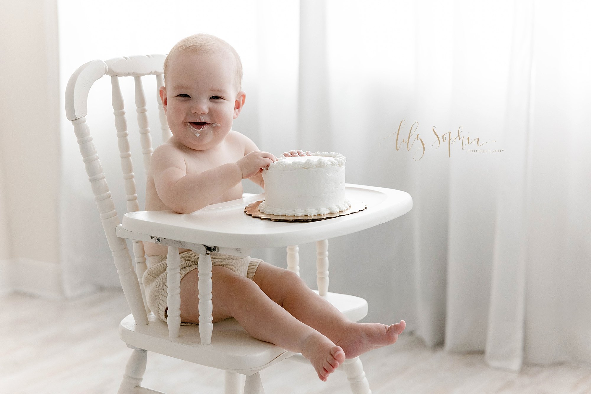  First birthday photo session of a one year old baby boy as he sits in an antique high chair with his cake on the tray and begins to taste the icing with natural light streaming in from a window behind him in the photography studio located in Ponce C