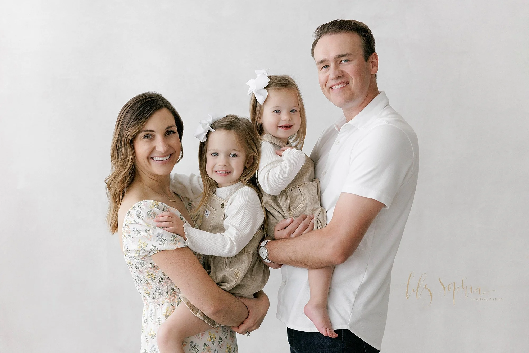  Family portrait of a mom holding her older daughter in her arms as she faces her husband holding their younger daughter in his arms with the girls’ backs touching taken near Brookhaven in Atlanta in a studio that uses natural light. 