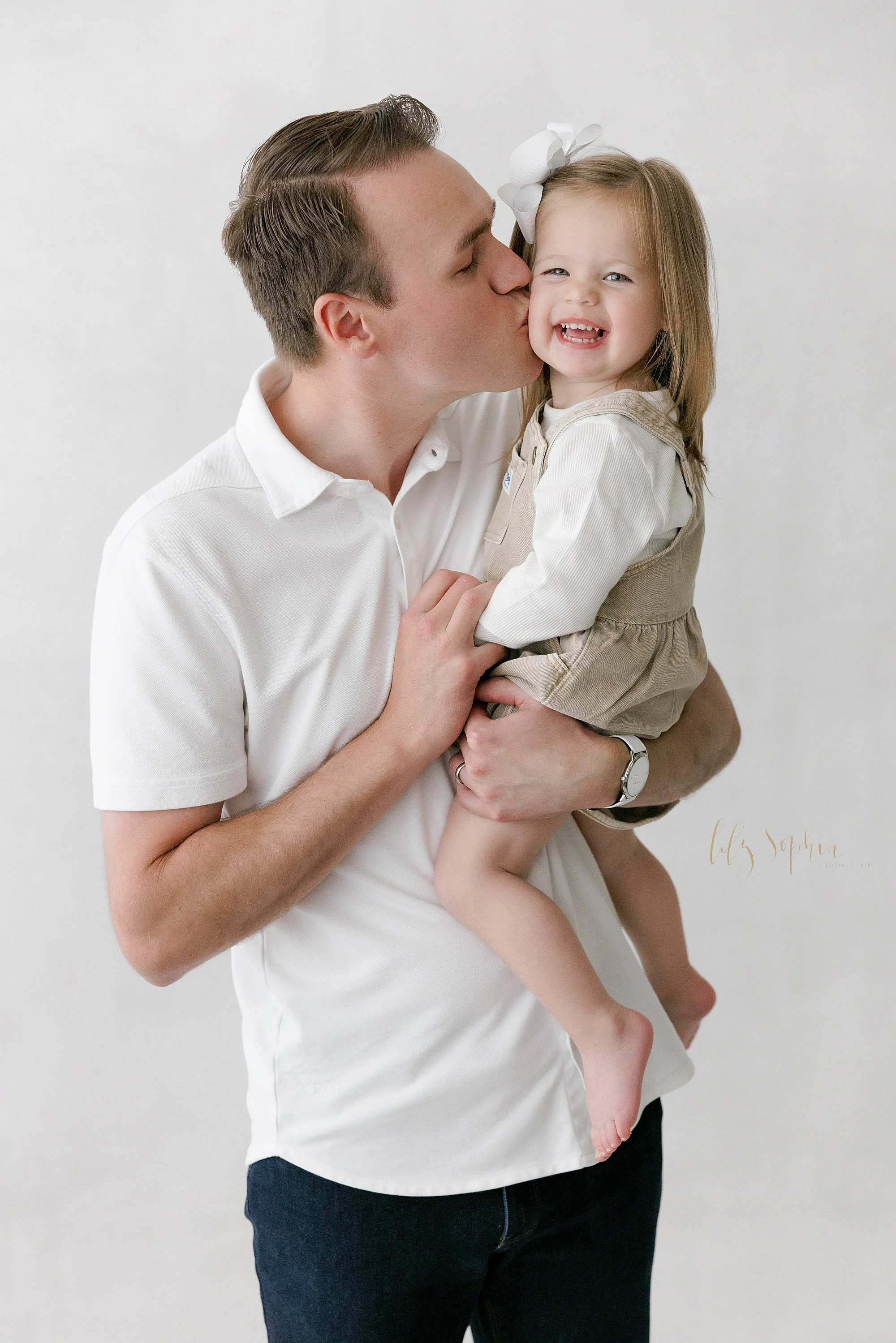  Family picture of a father holding his young daughter in his arms as he kisses her on the cheek taken in natural light near Old Fourth Ward in Atlanta in a photography studio. 