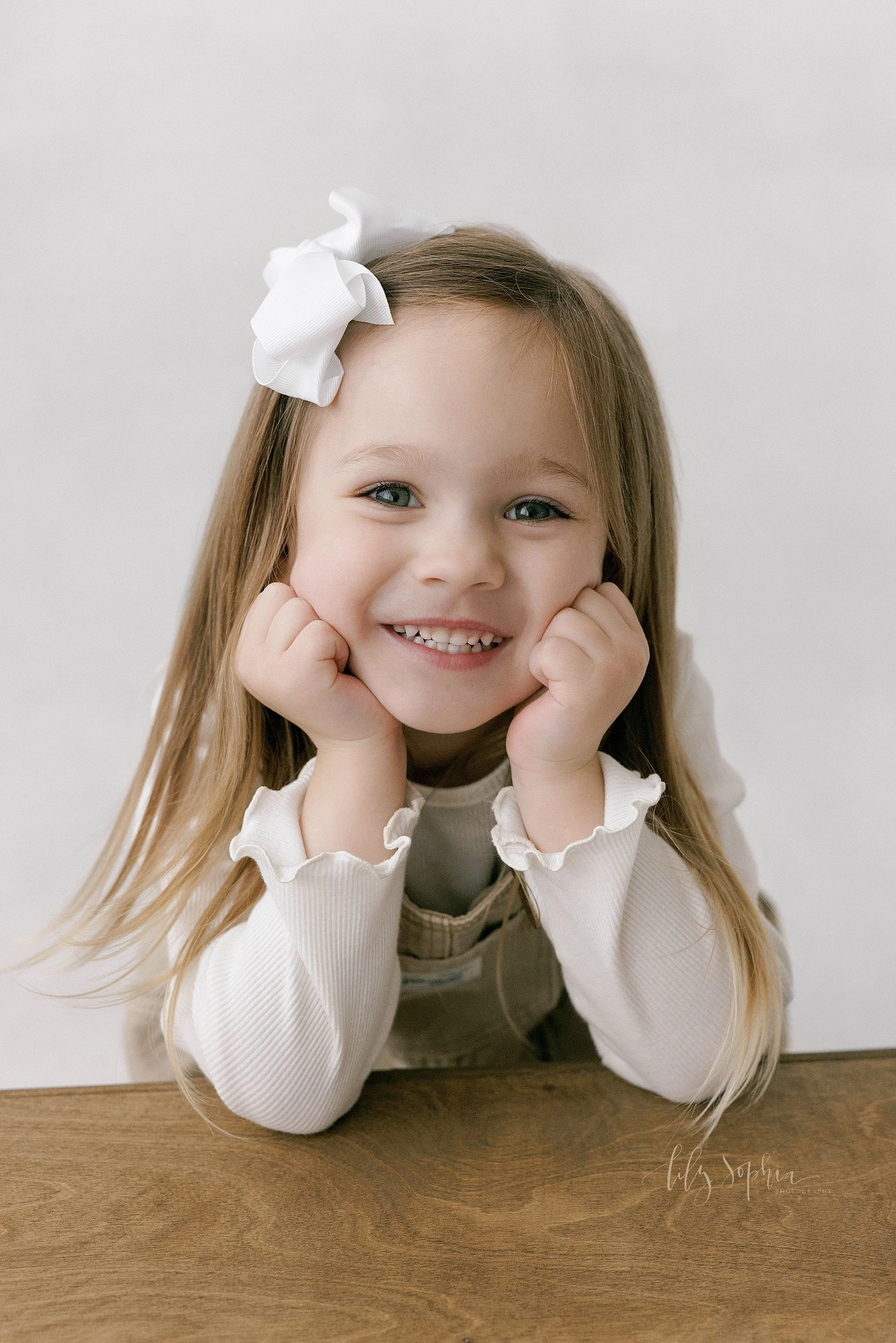  Close-up portrait of a smiling young girl as she leans her elbows on a wooden box and places her fists on her cheeks during a family photography session in a natural light studio near Midtown in Atlanta, Georgia. 