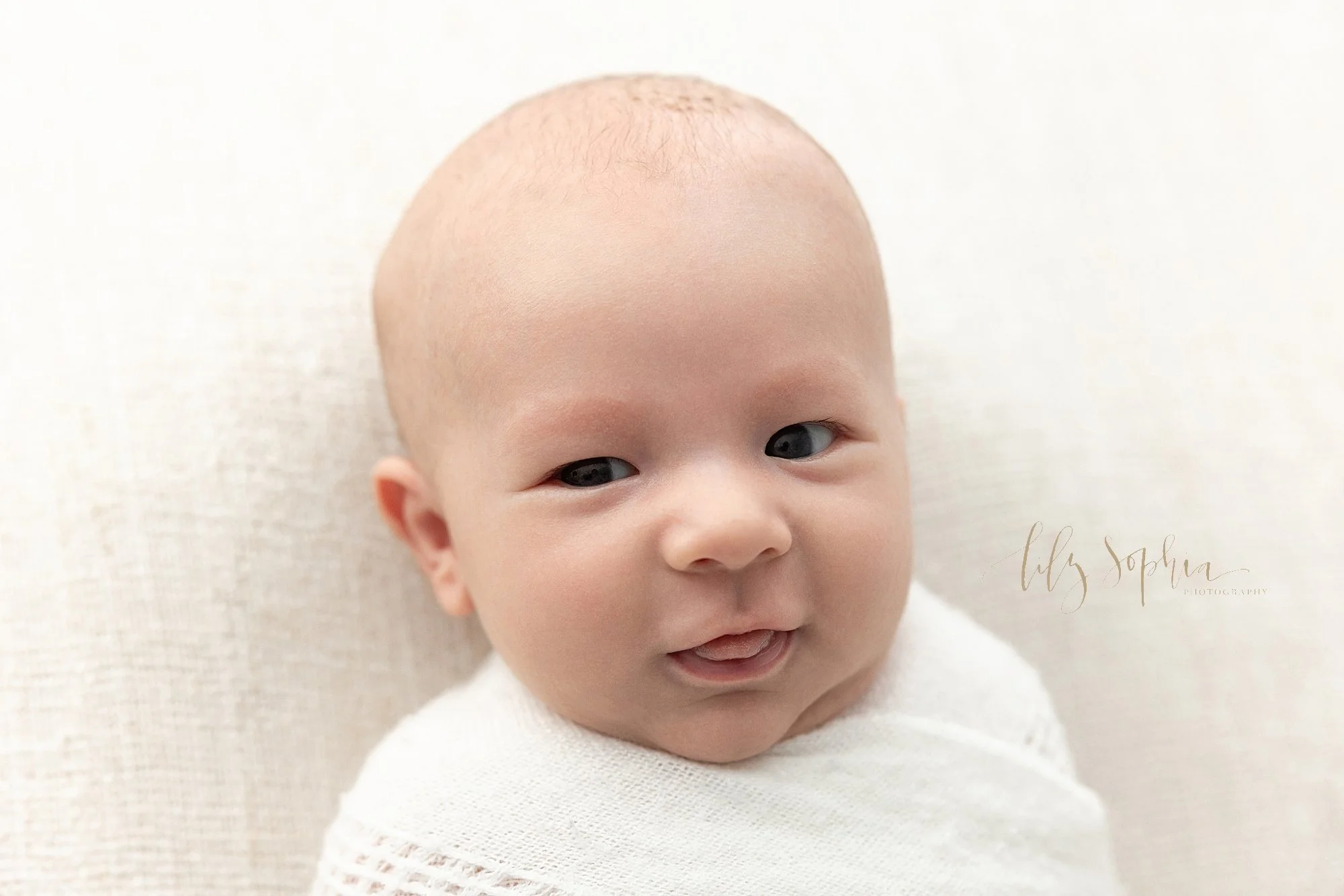  Close-up newborn portrait of an awake baby boy as he sticks out his tongue while lying on his back in a photography studio near Alpharetta in Atlanta that uses natural light. 