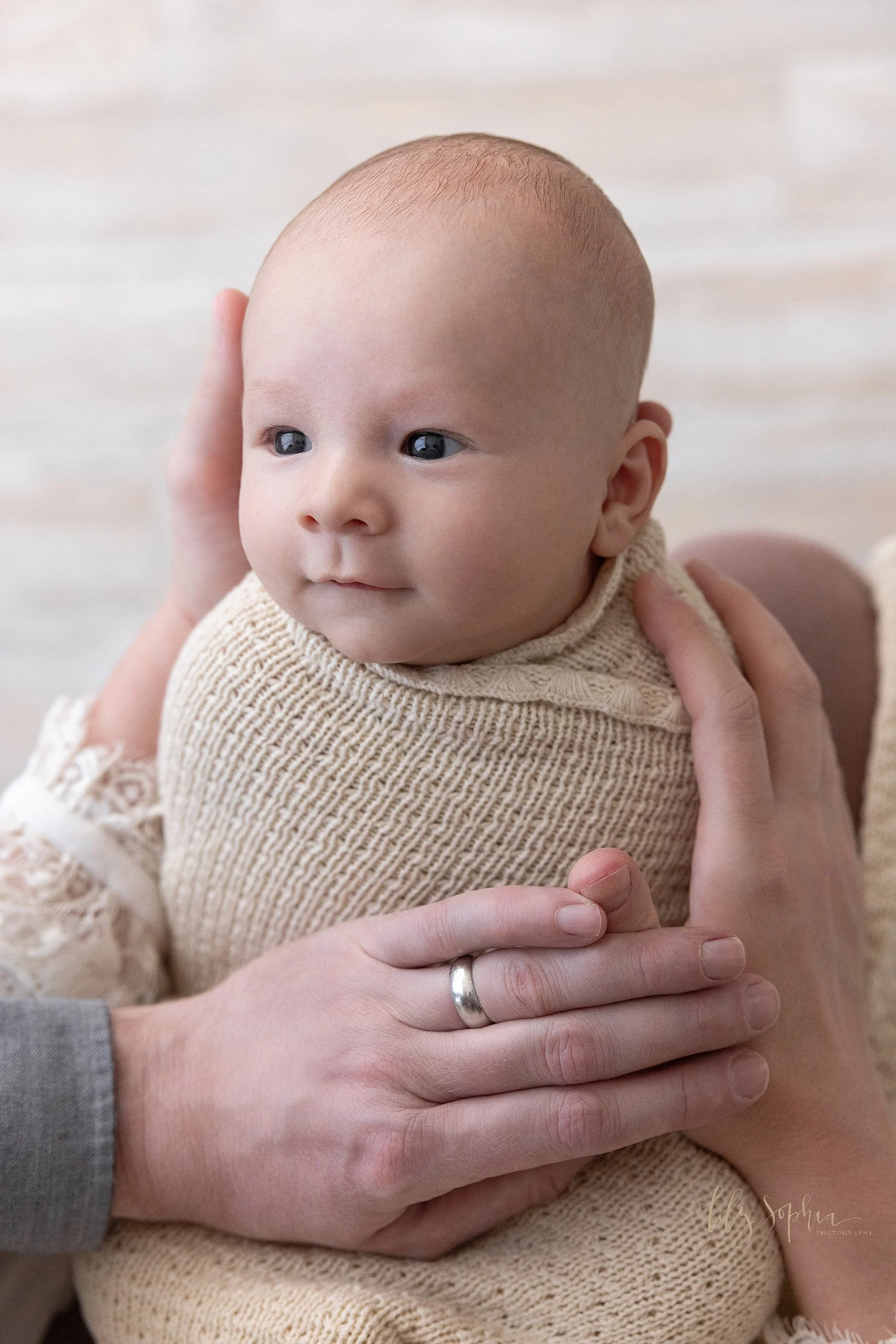  Newborn photo session with a newborn baby boy as his mom holds his head in her hands and dad places his hand on his body while the baby boy looks at his father taken using natural light near Ansley Park in Atlanta, Georgia in a photography studio. 