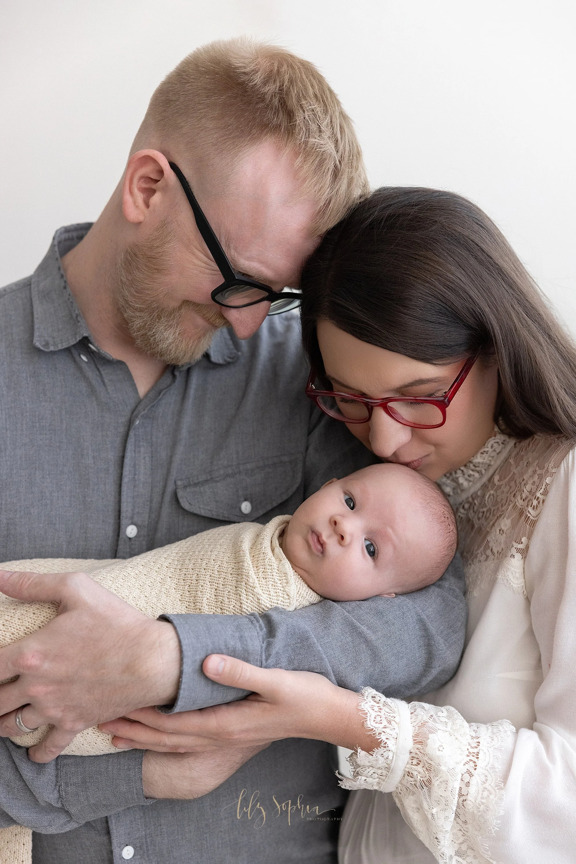  Dad holds his awake newborn baby boy in his right arm as his wife stands next to him on his right and kisses the crown of her son and her husband smiles as he watches the interaction taken in Ponce City Market  in Atlanta in a photography studio tha