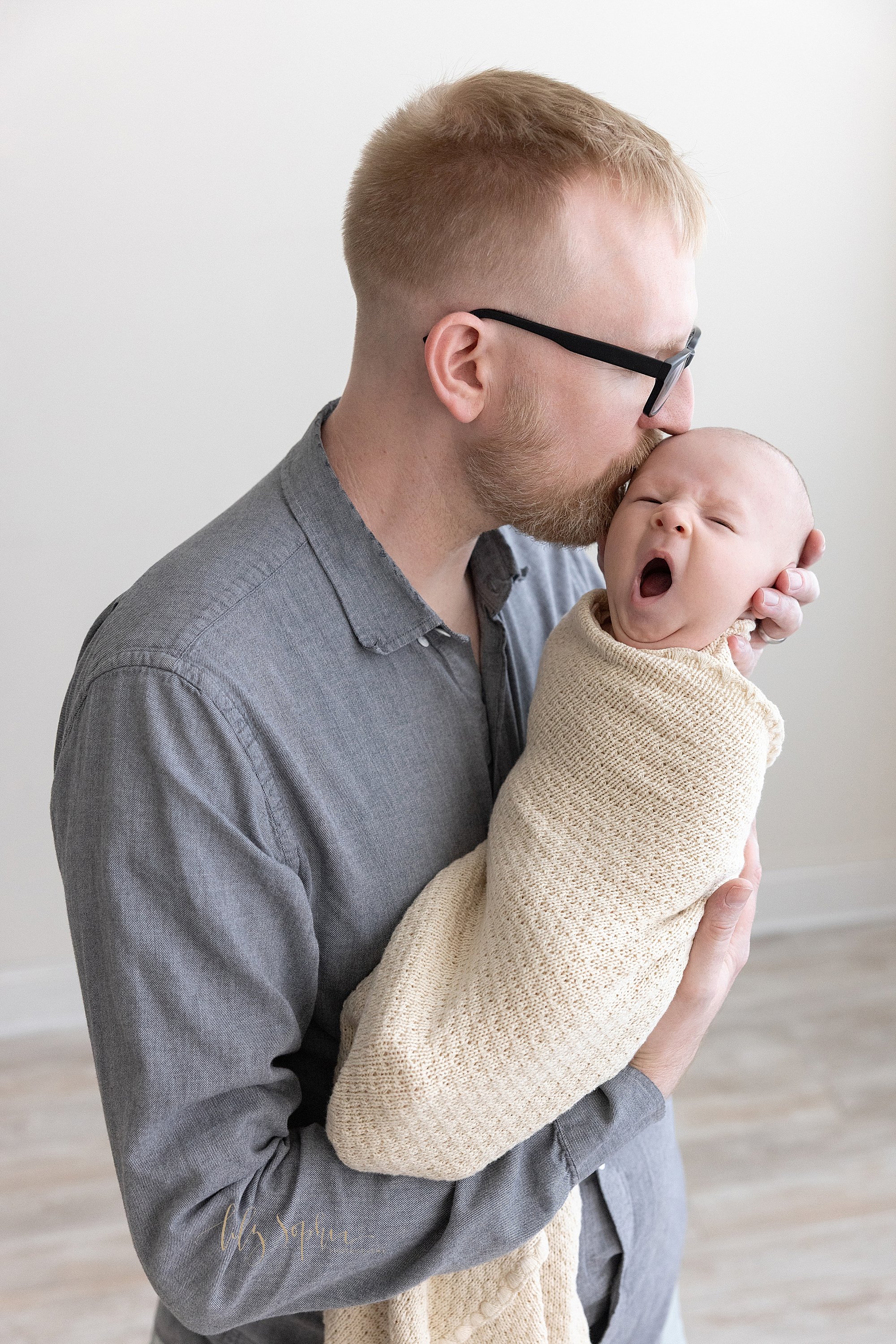  A father stands in a photography studio holding his yawning newborn baby boy as he kisses his son’s head taken in natural light near Roswell in Atlanta. 