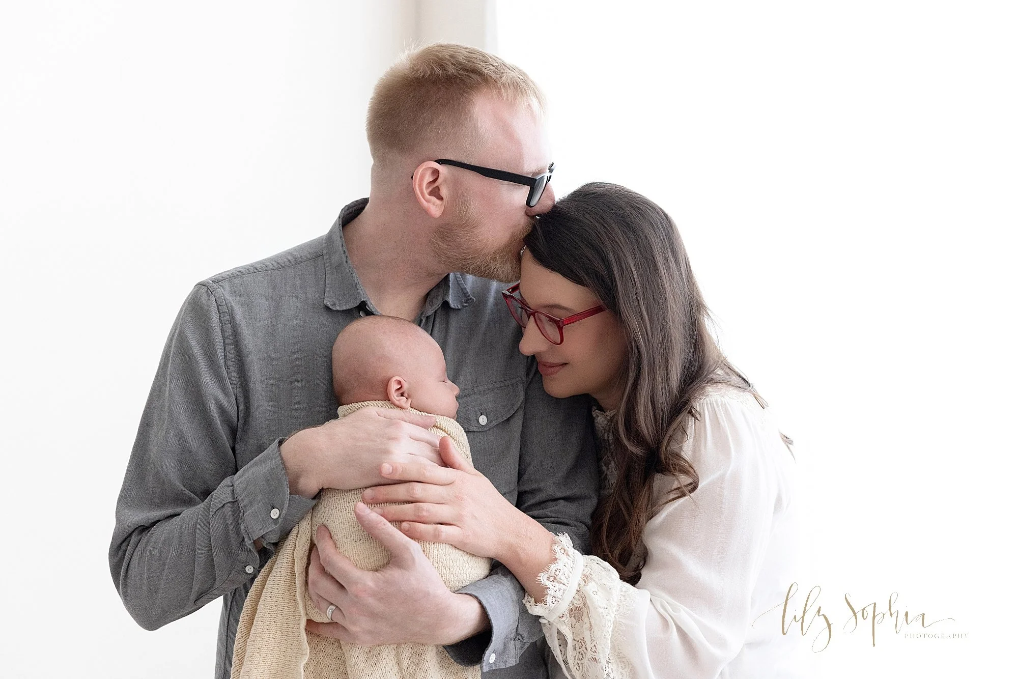  Family newborn photo of dad holding his peacefully sleeping baby boy against his chest as mom stands to dad’s right side and looks at her son while placing her left hand on him and dad kisses his wife’s head while the couple stand next to a window s