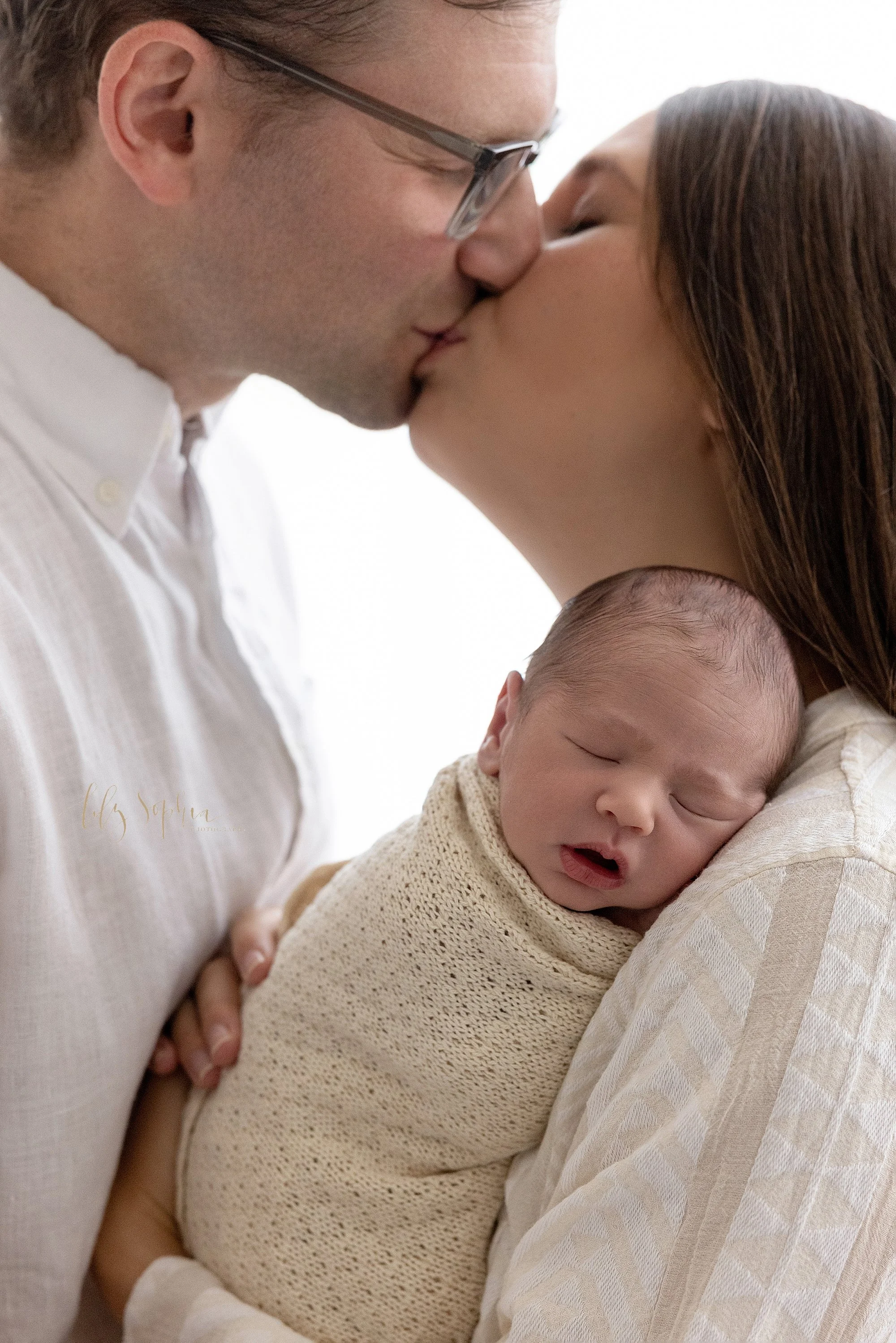  As this newborn baby boy sleeps on his mom’s left shoulder, the parents kiss one another while standing in front of a window streaming natural light in a photography studio located near Ansley Park in Atlanta. 