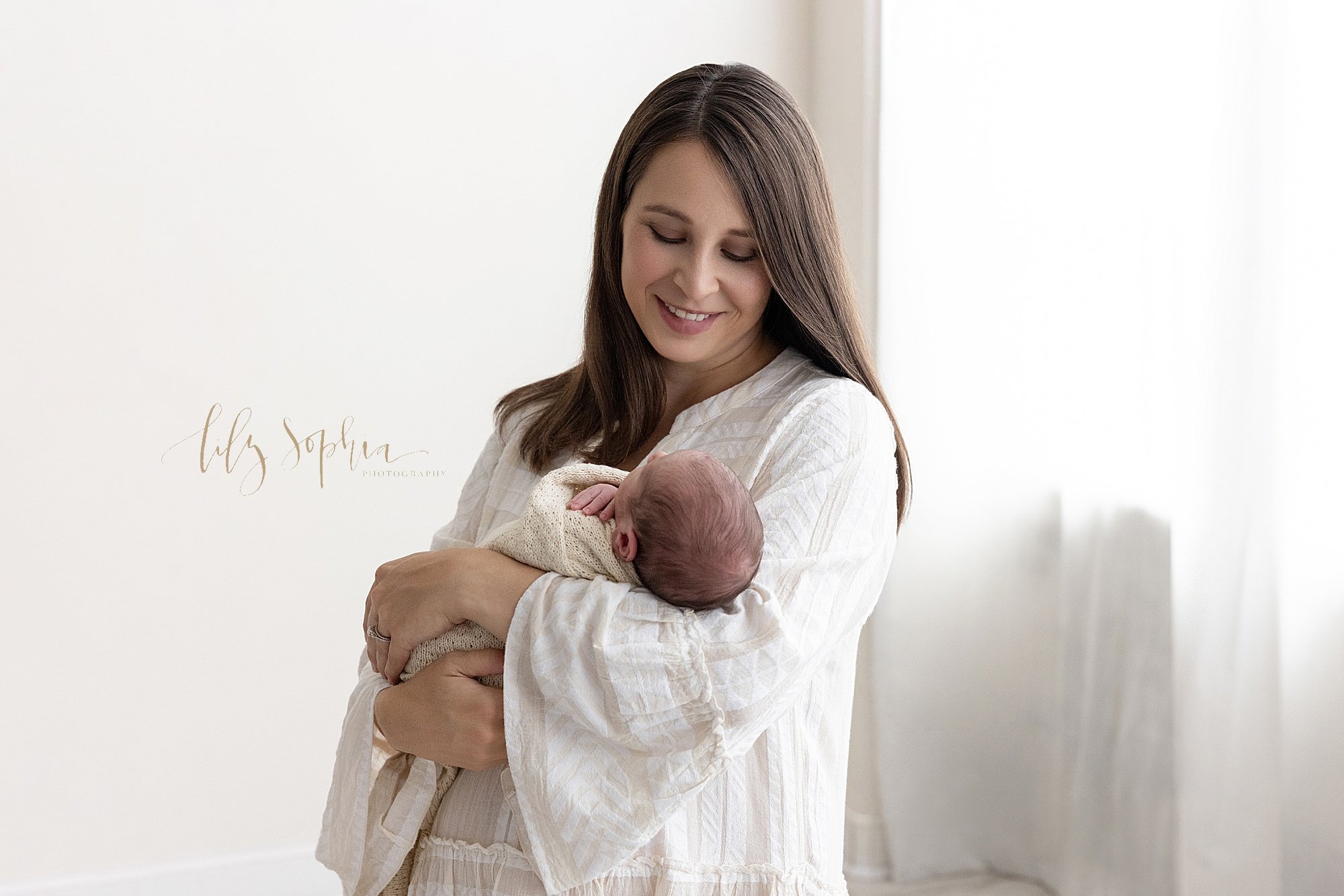  The joy and admiration of a mother for her newborn baby boy is captured in this photo as mom cradles her son in her left arm while standing next to a window streaming natural light in a photography studio located near Buckhead in Atlanta, Georgia. 