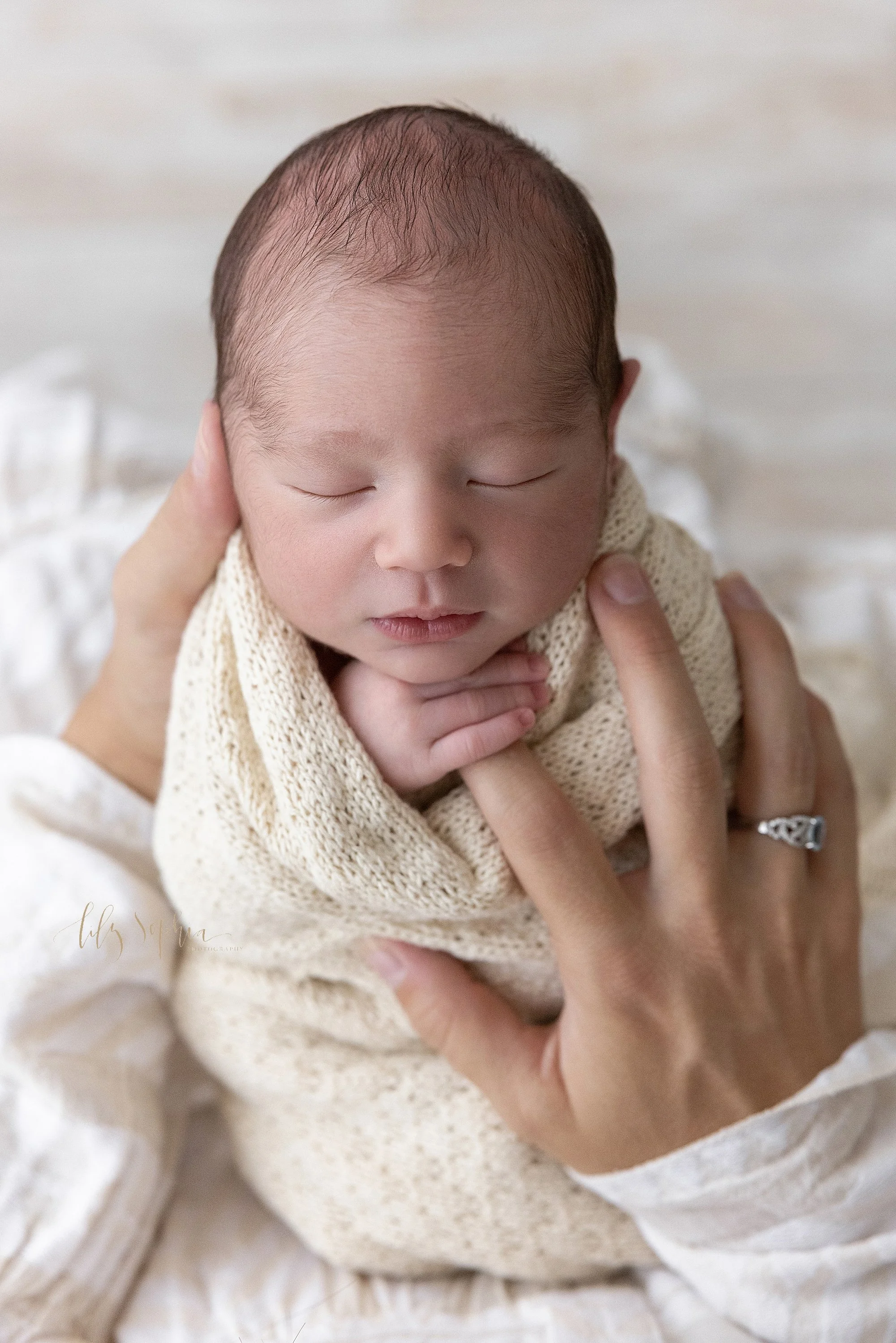  Close-up newborn photo of a peacefully sleeping newborn baby boy as he holds the index finger of his mother’s right hand taken using natural light near Morningside in Atlanta in a photography studio. 