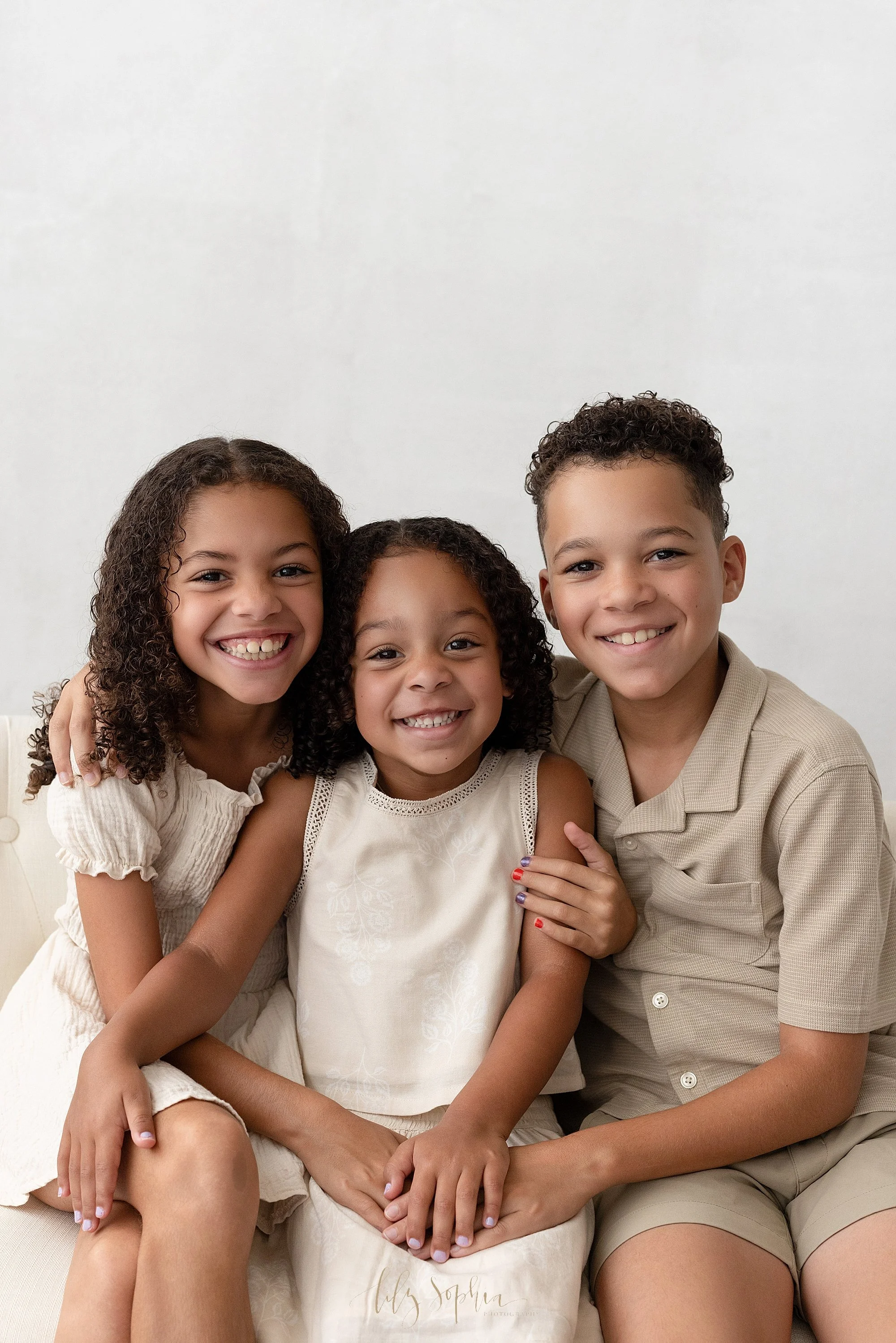  Family sibling portrait with the older sister on one side of her younger sister and the older brother on the other side as he puts his hand around his sister’s shoulder and she puts her hand around her younger sister’s shoulder with all of them plac