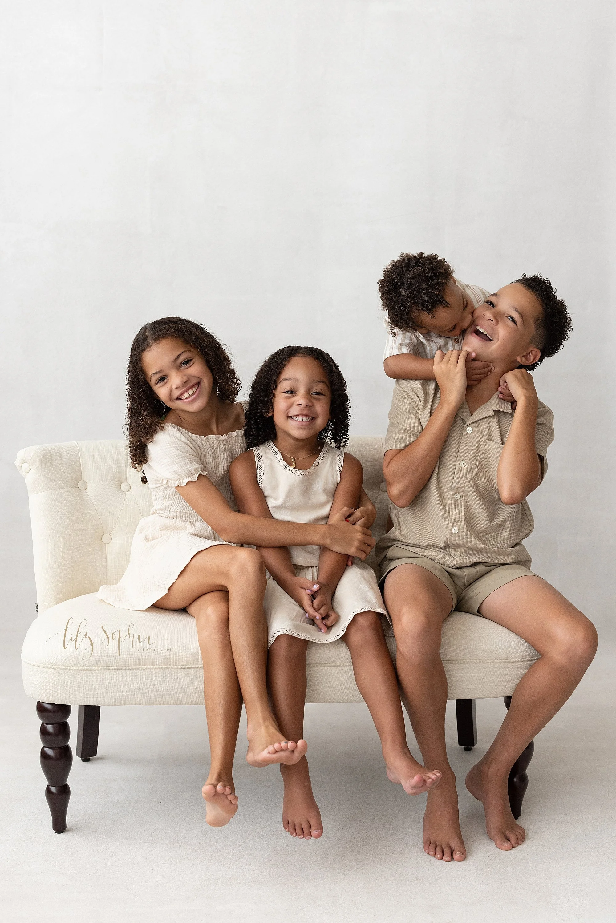  Sibling photo during a family photography session featuring two sisters with the older sister wrapping her arms around her younger sister as they sit on a tufted settee next to their brothers with the younger brother standing behind his older brothe