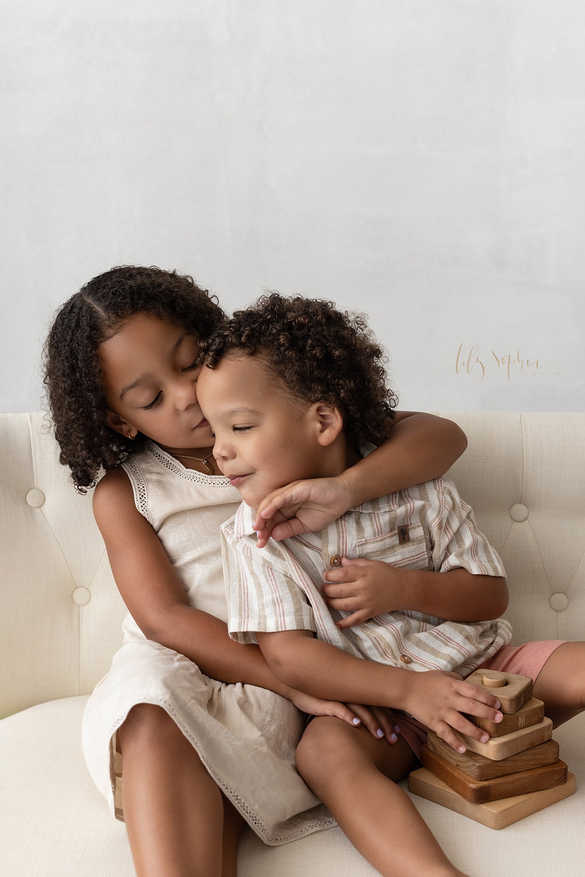  The love between siblings is found as this older sister sits on a settee with her younger brother as he plays with a wooden stacking toy and she pulls his head toward her to kiss him on the cheek during a family photo shoot near Old Fourth Ward in A