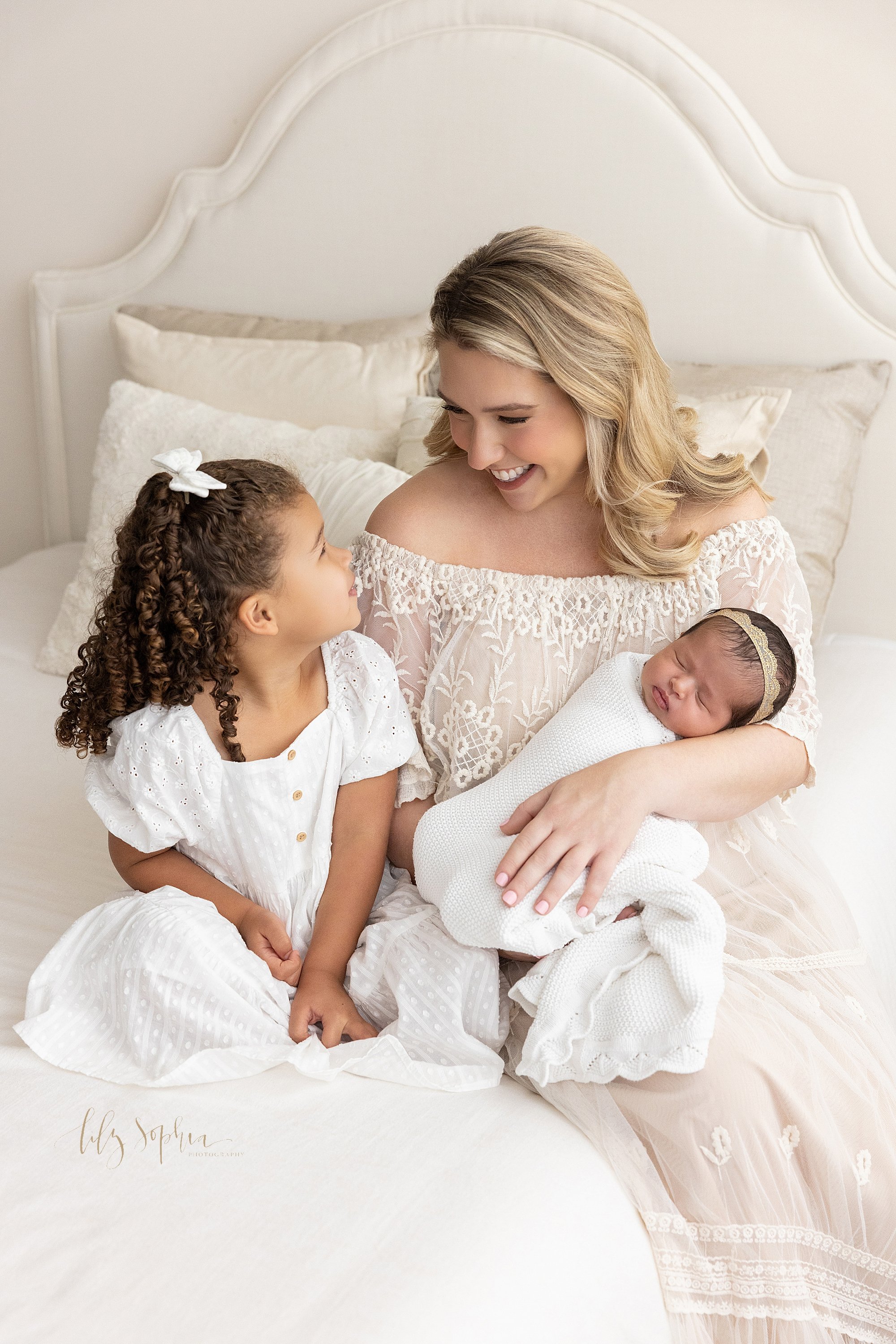  Family newborn picture of a mother holding her newborn baby daughter in her left arm as her older daughter sits next to her on a bed and the two talk with one another taken near Ansley Park in Atlanta in a photography studio that uses natural light.