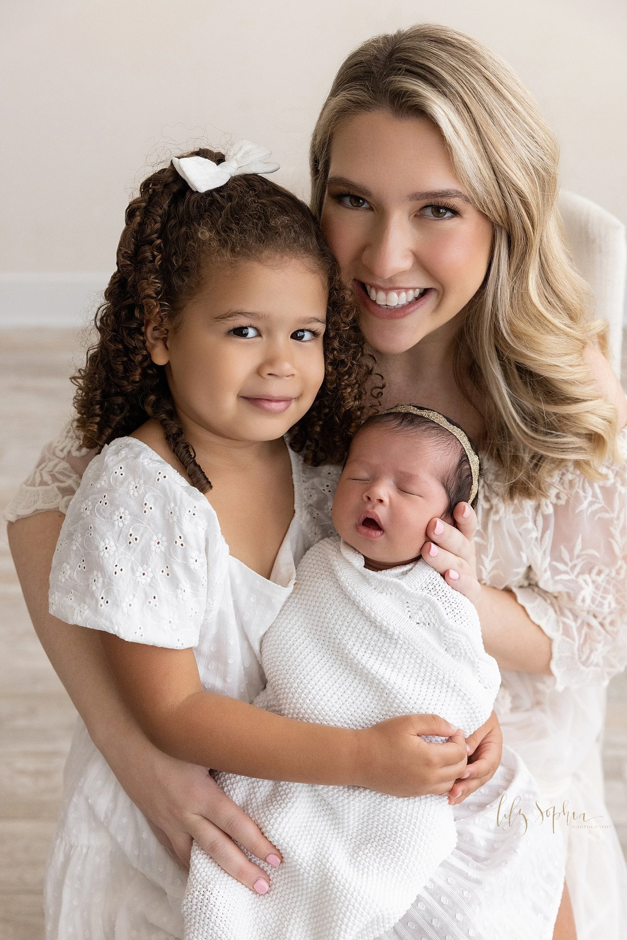  Newborn family portrait of a mother with her young daughter on her knee as she helps her daughter hold her newborn baby sister on her lap taken near Midtown in Atlanta, Georgia in a natural light studio. 