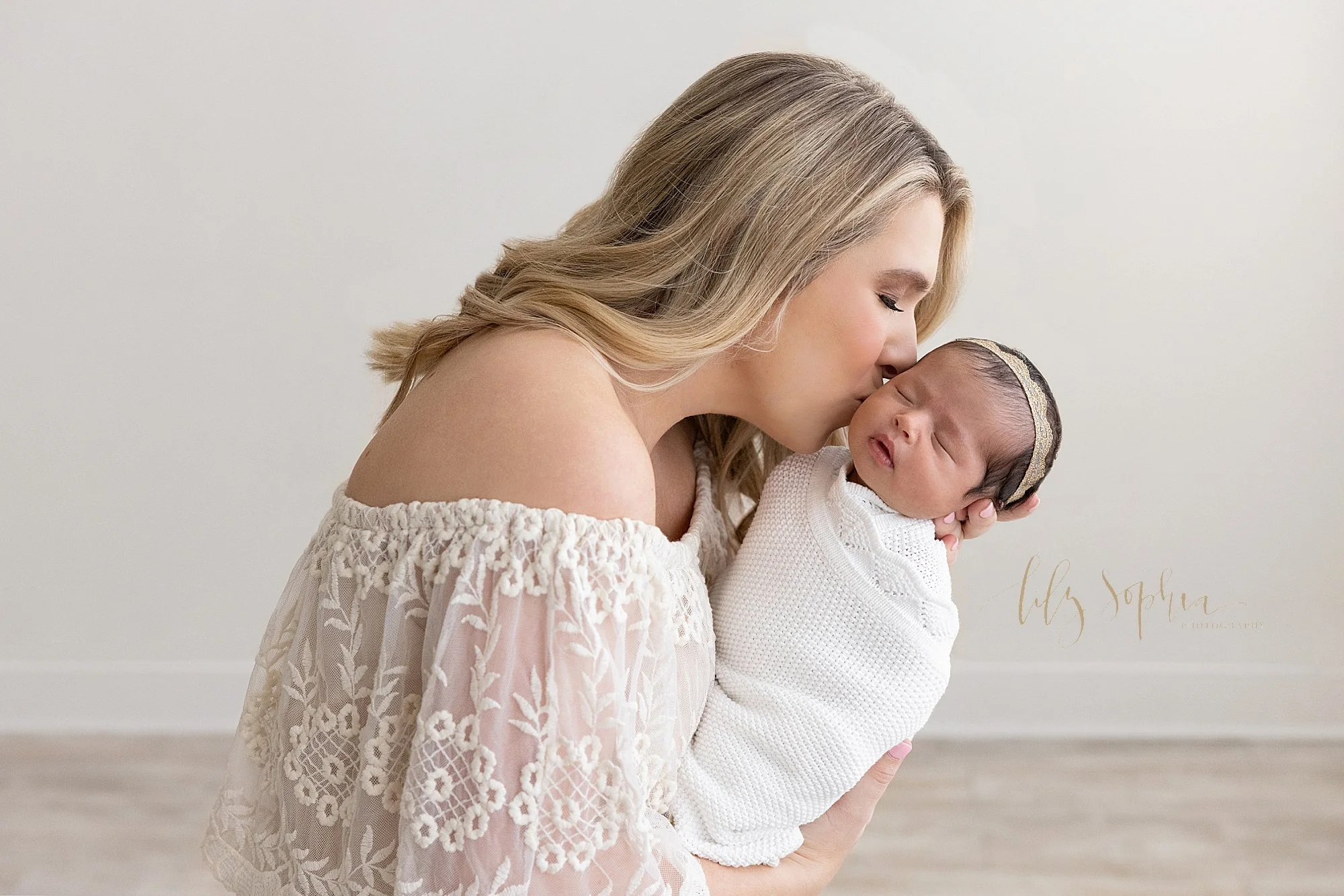  The love a mother has for her newborn daughter is captured during this newborn photography session in a natural light studio as mom holds her daughter in her left hand and kisses her cheek as she peacefully sleeps taken near Oakhurst in Atlanta, Geo