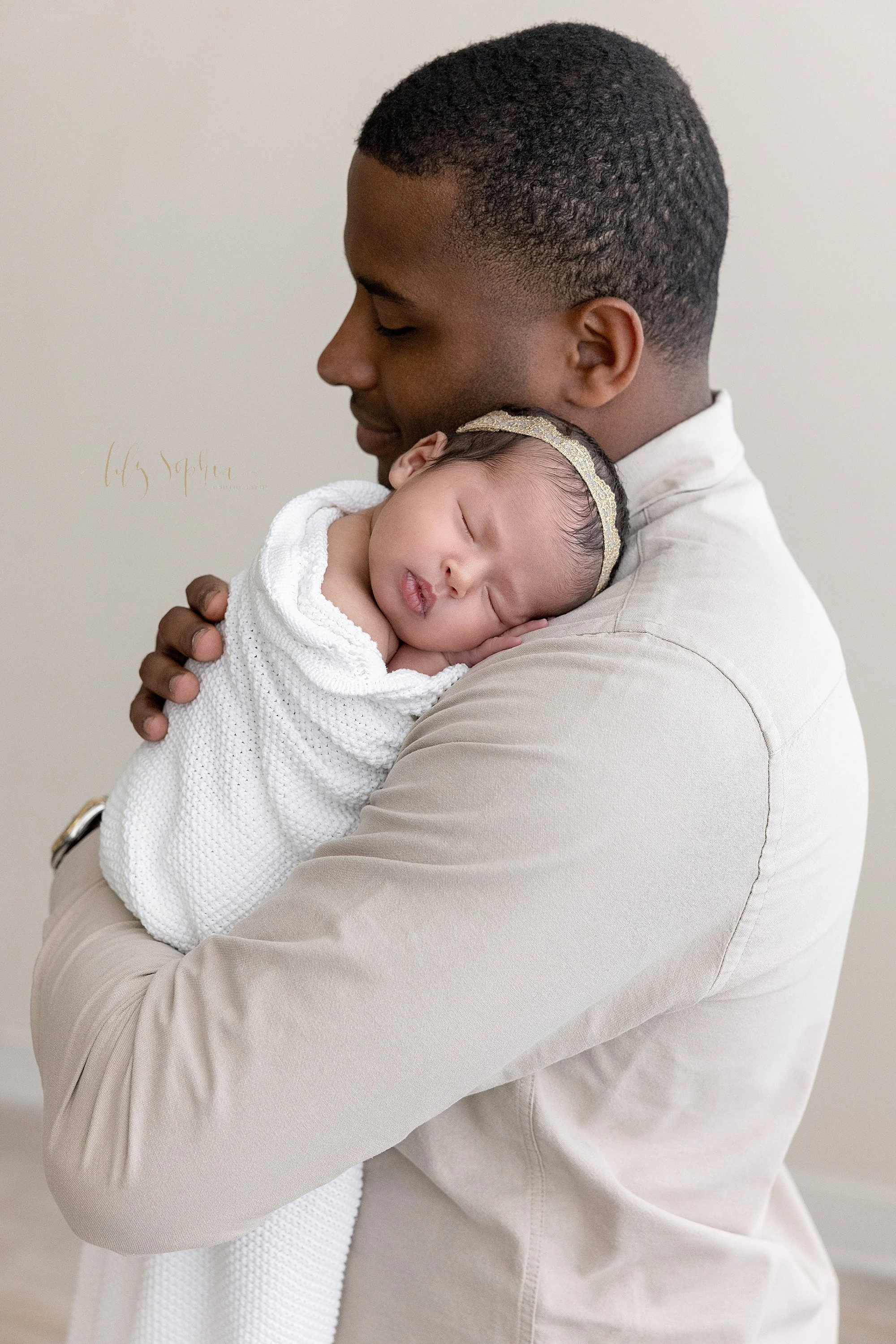  A treasured moment between a father and his newborn baby girl as she sleeps on his left shoulder while he stands in a photography studio near Buckhead in Atlanta that uses natural light. 
