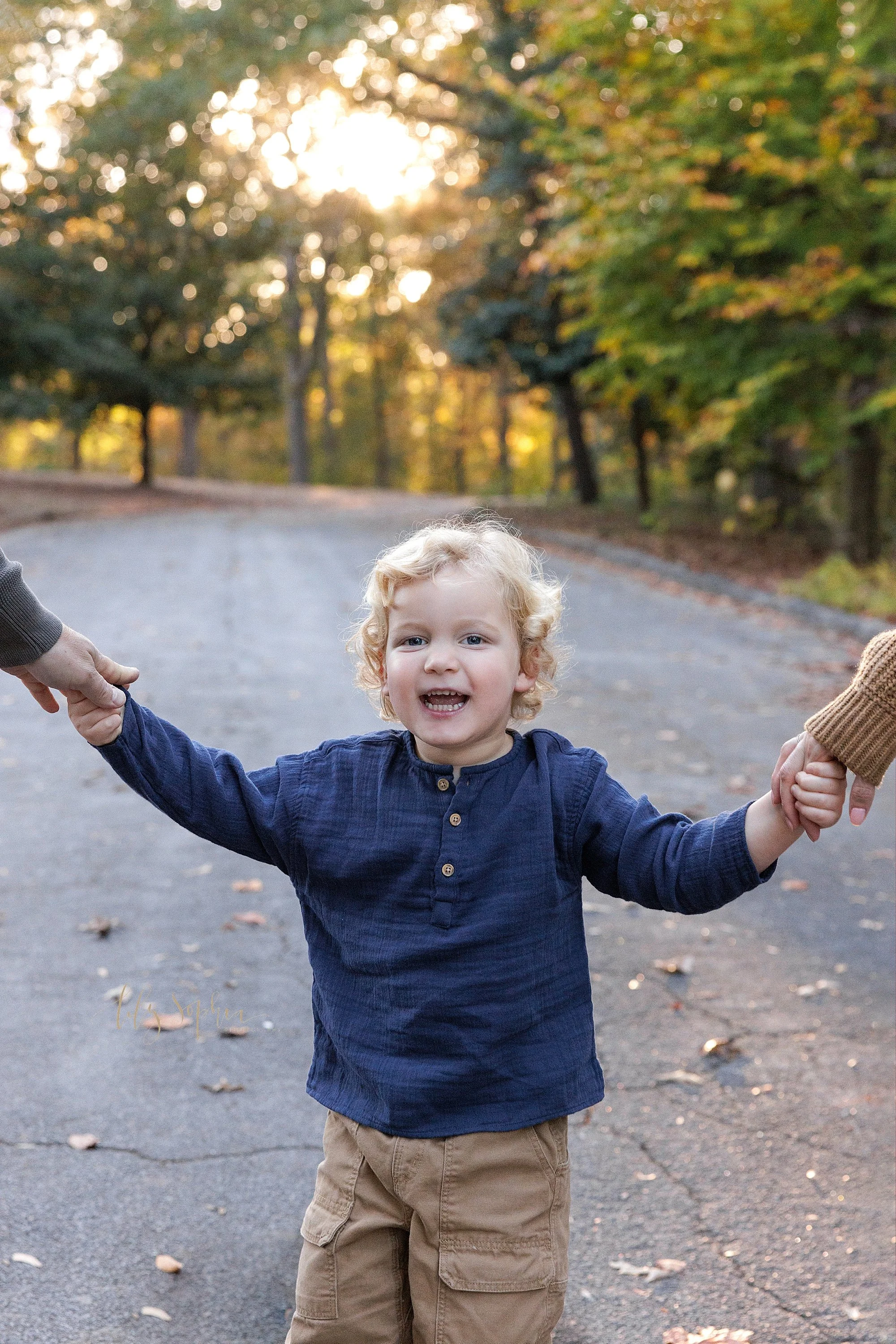  The pure joy and security of this toddler boy is captured as he walks along an Atlanta park walkway holding his parents’ hands during sunset in autumn. 
