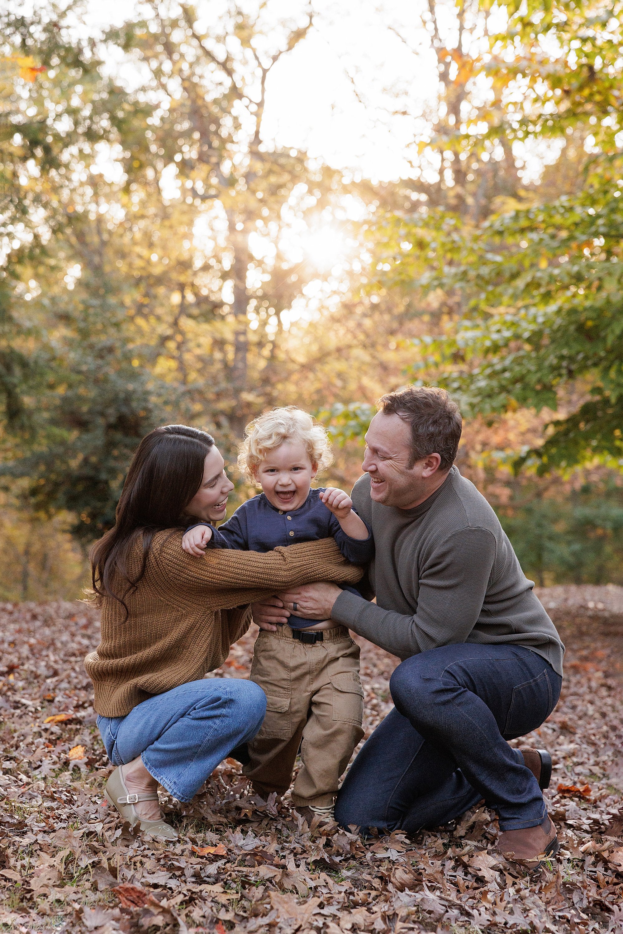  Family photo session in a park near Atlanta with a Mom and Dad  catching their toddler son as he tries to run from them during autumn at sunset. 