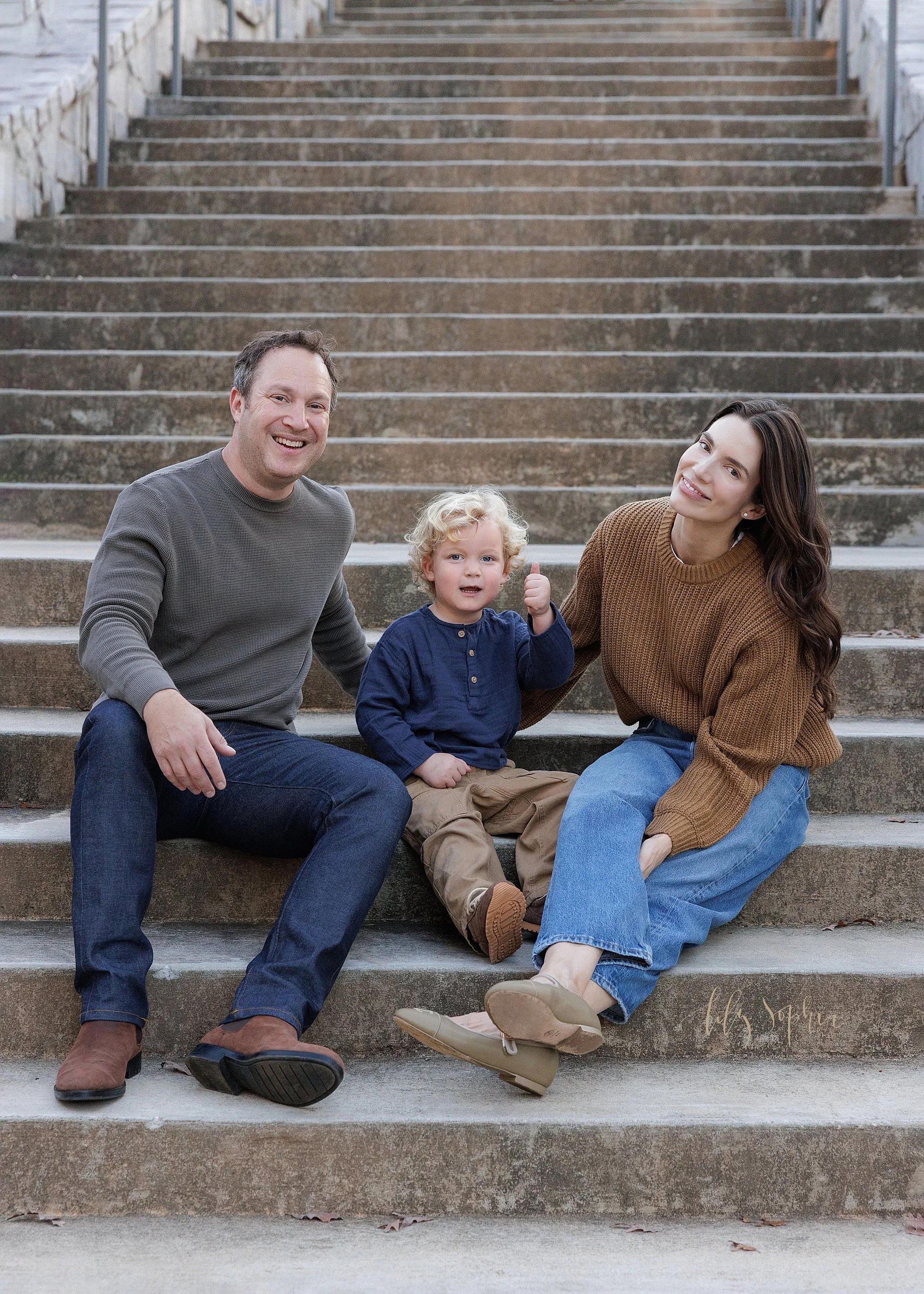  A family sits at the base of a stone staircase in an Atlanta park with their toddler son between them as he gives a thumbs up taken during autumn at sunset for a family photography session. 