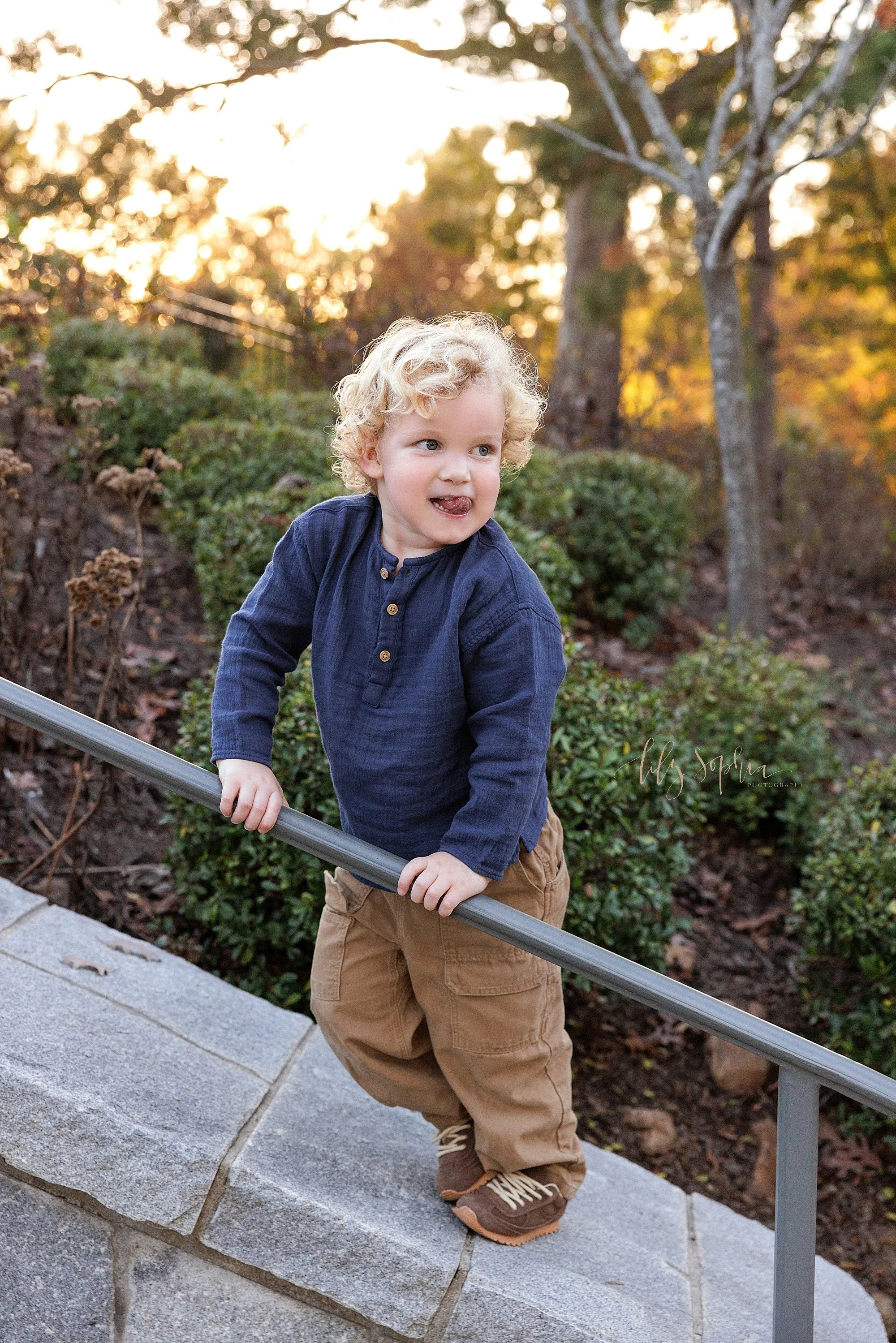  The determination of a toddler boy with his tongue sticking out as he walks up a stone wall while holding on to the railing in a park near Atlanta, Georgia at sunset during autumn for a family photo shoot. 