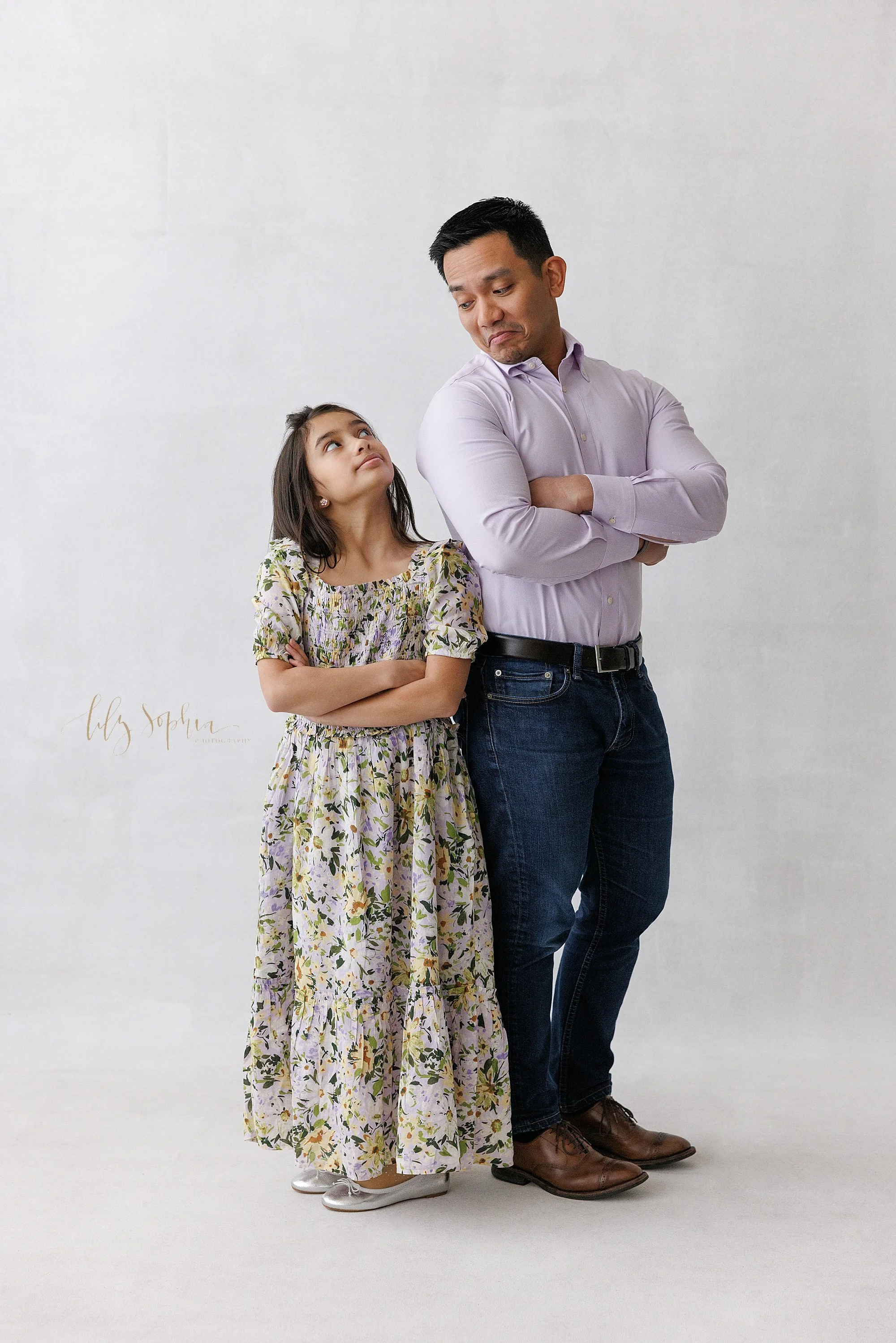  A father and daughter stand with their hips together and their arms folded in front of them as dad looks over his right shoulder down at his daughter who is looking up at him during a family photo shoot in a photography studio located near Cumming i