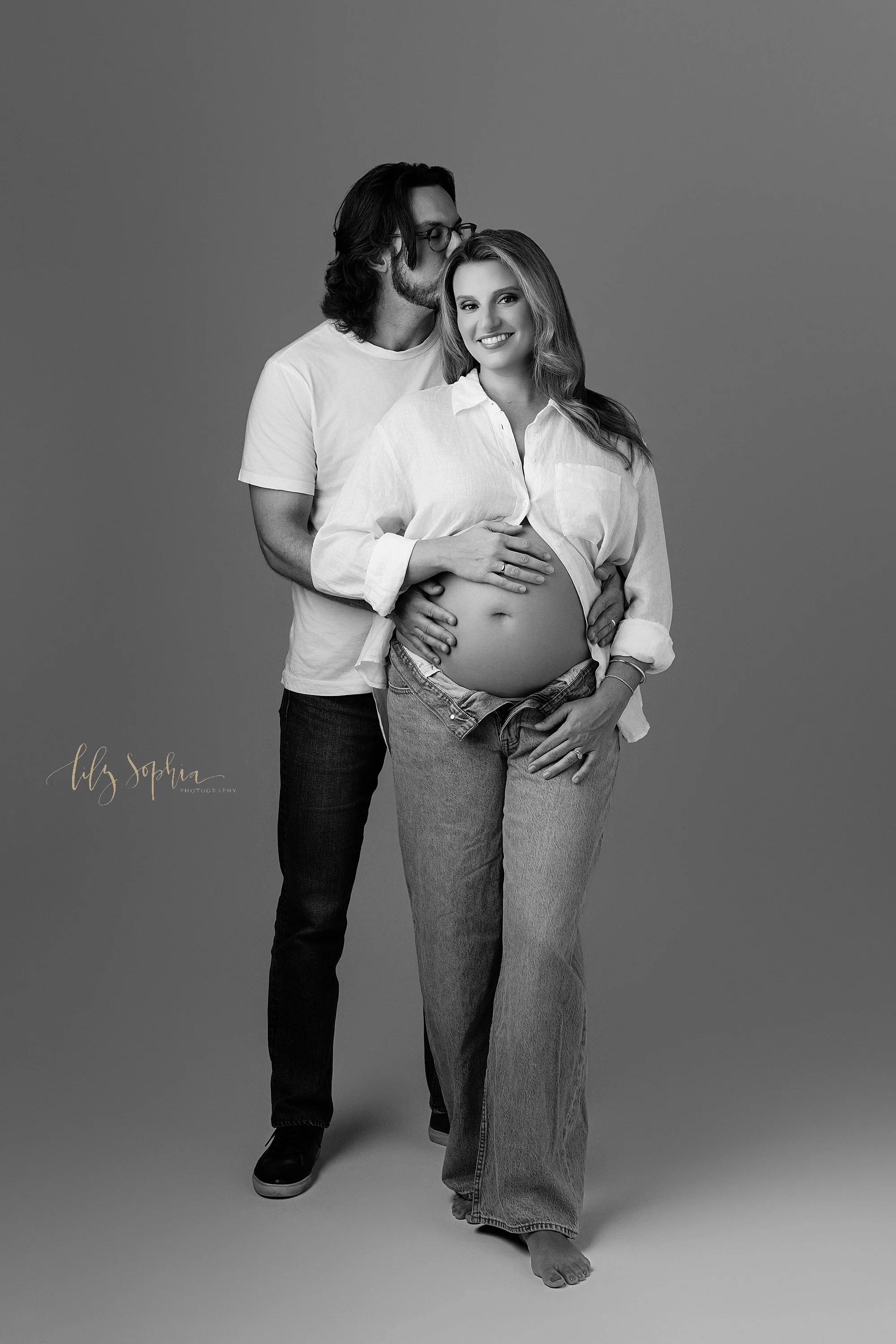  Modern maternity photo session with an expectant couple standing in a studio with the mother wearing unzipped jeans and a buttoned down white collared shirt that is open to expose her belly as the father stands behind her and places his hands on the