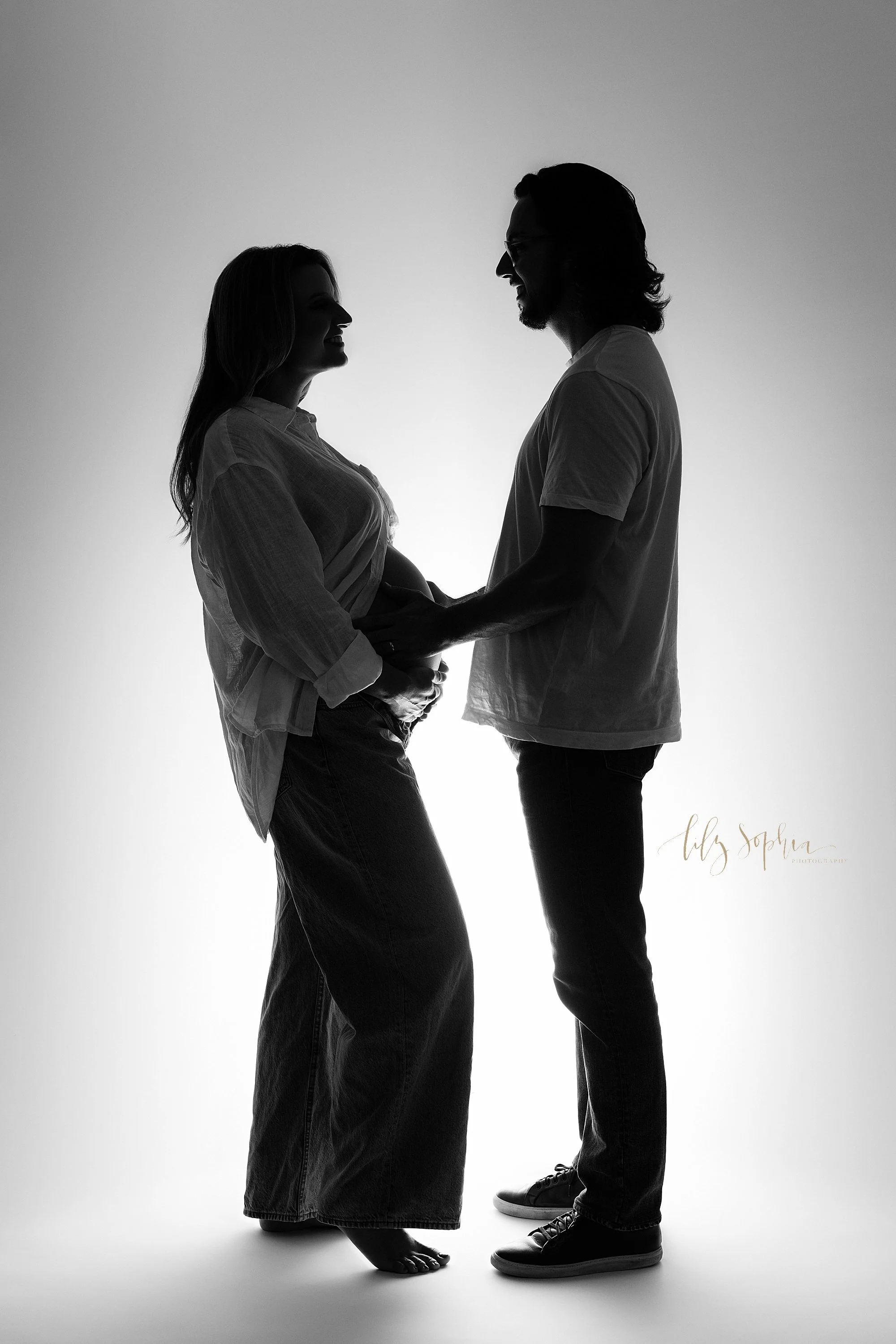  A modern maternity photo session in a photography studio with the expectant couple facing one another with mom holding the base of her belly and dad placing his hands on their child in utero taken near Decatur in Atlanta. 