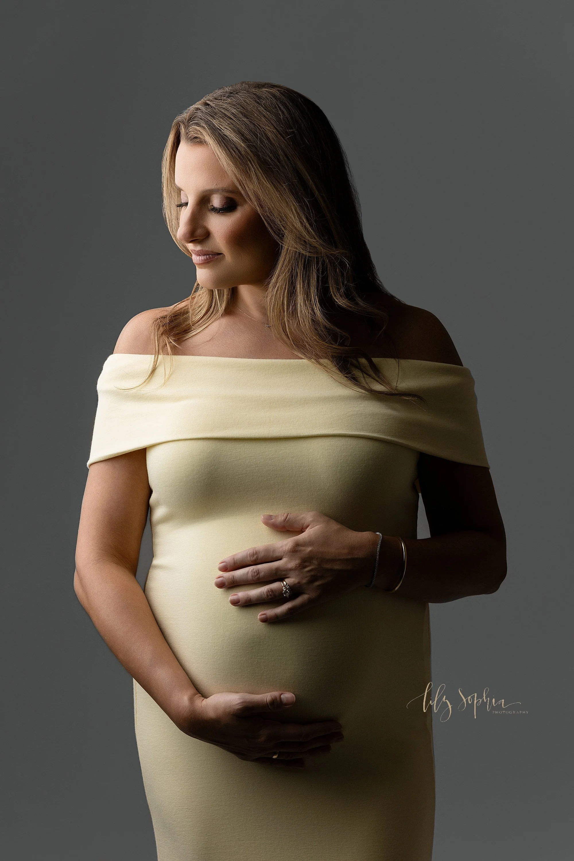  A pregnant mother stands wearing a knit off the shoulder sleeveless gown and she places her hands on her child in utero as she closes her eyes and looks toward her right shoulder contemplating the upcoming birth of her child taken in a photography s