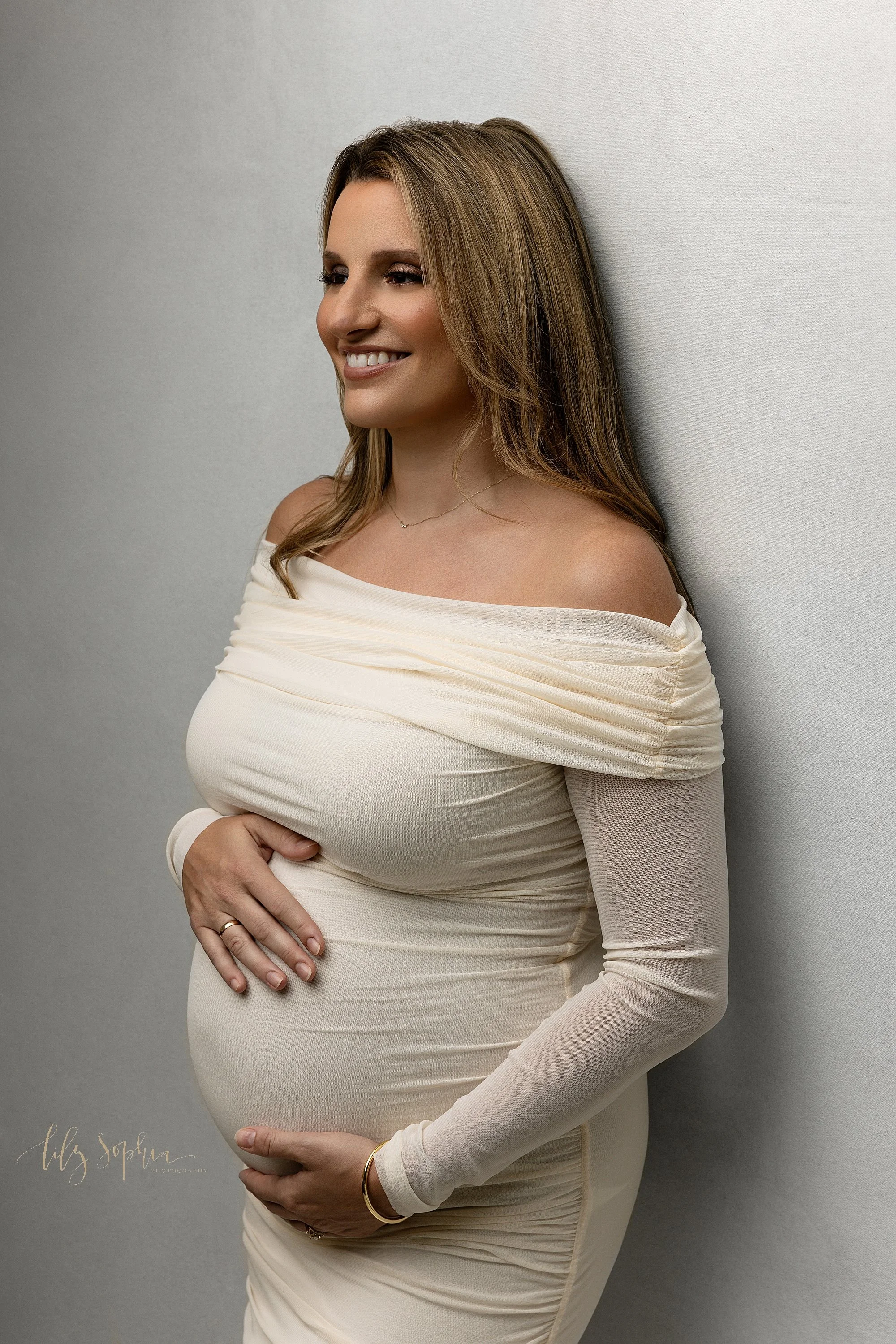  An expectant mother stands with her back to a wall wearing a knit off the shoulder long-sleeved gown as she frames her belly with her hands during a maternity session in a studio located near Brookhaven in Atlanta, Georgia. 