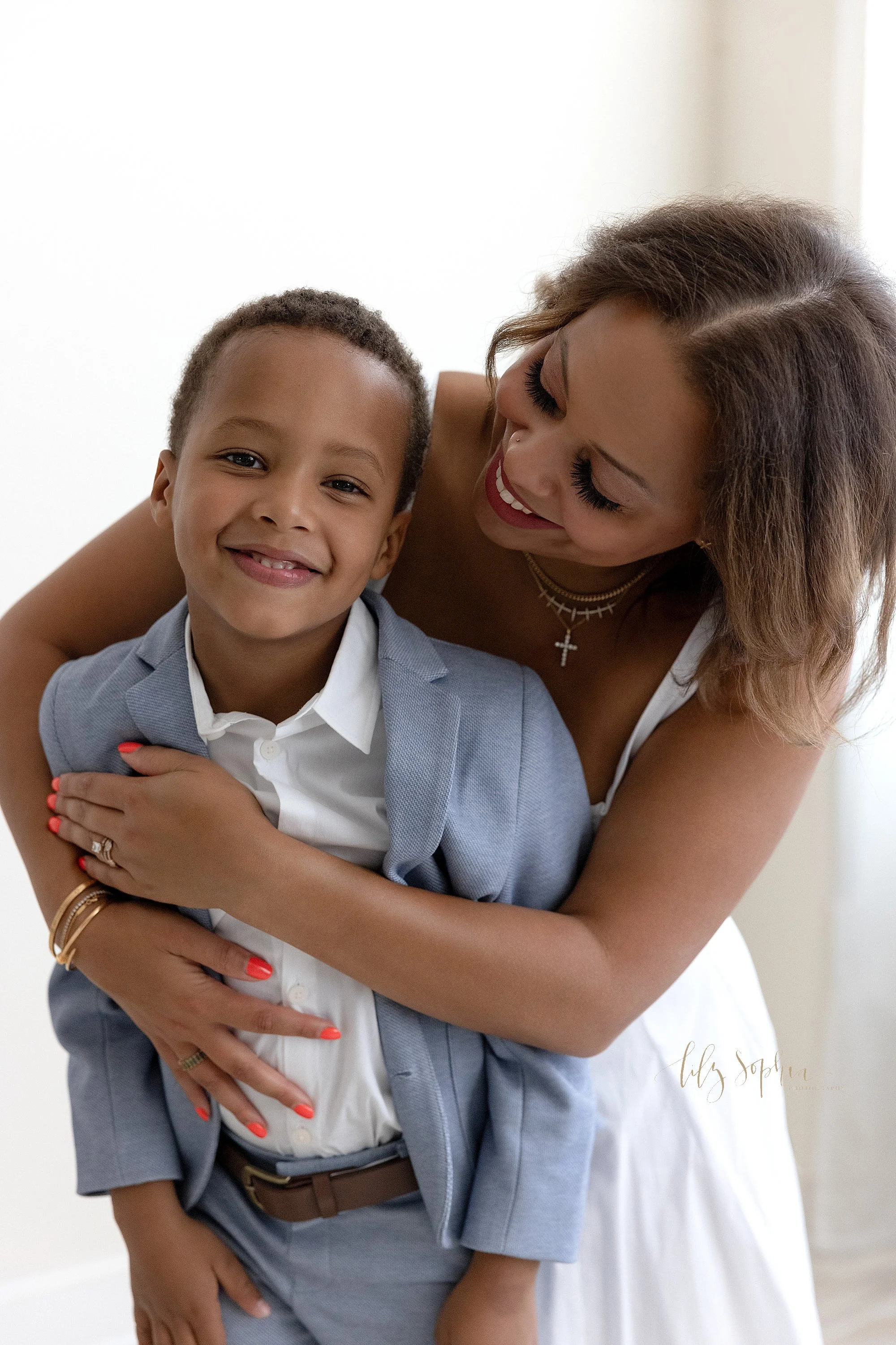  The love of a black mom for her son is captured in a photography studio using natural light as mom wraps her arms around her son from behind him taken near Kirkwood in Atlanta during a family photo shoot. 