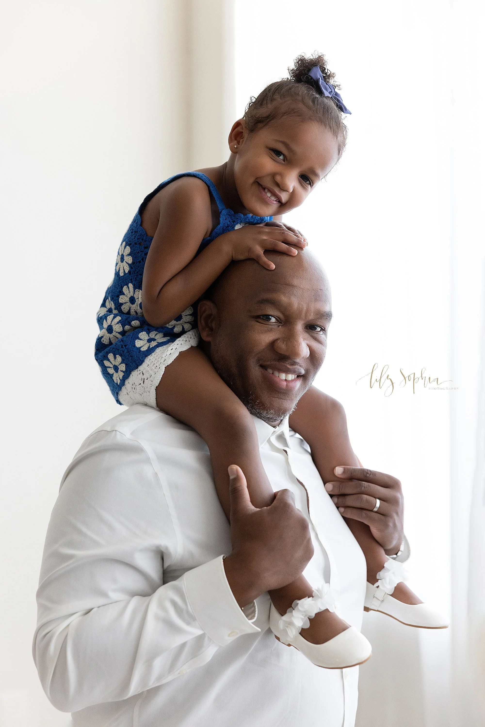  An African-American father holds his young daughter on his shoulders during a family photo session in a studio as he stands next to window streaming natural light in Midtown in Atlanta. 