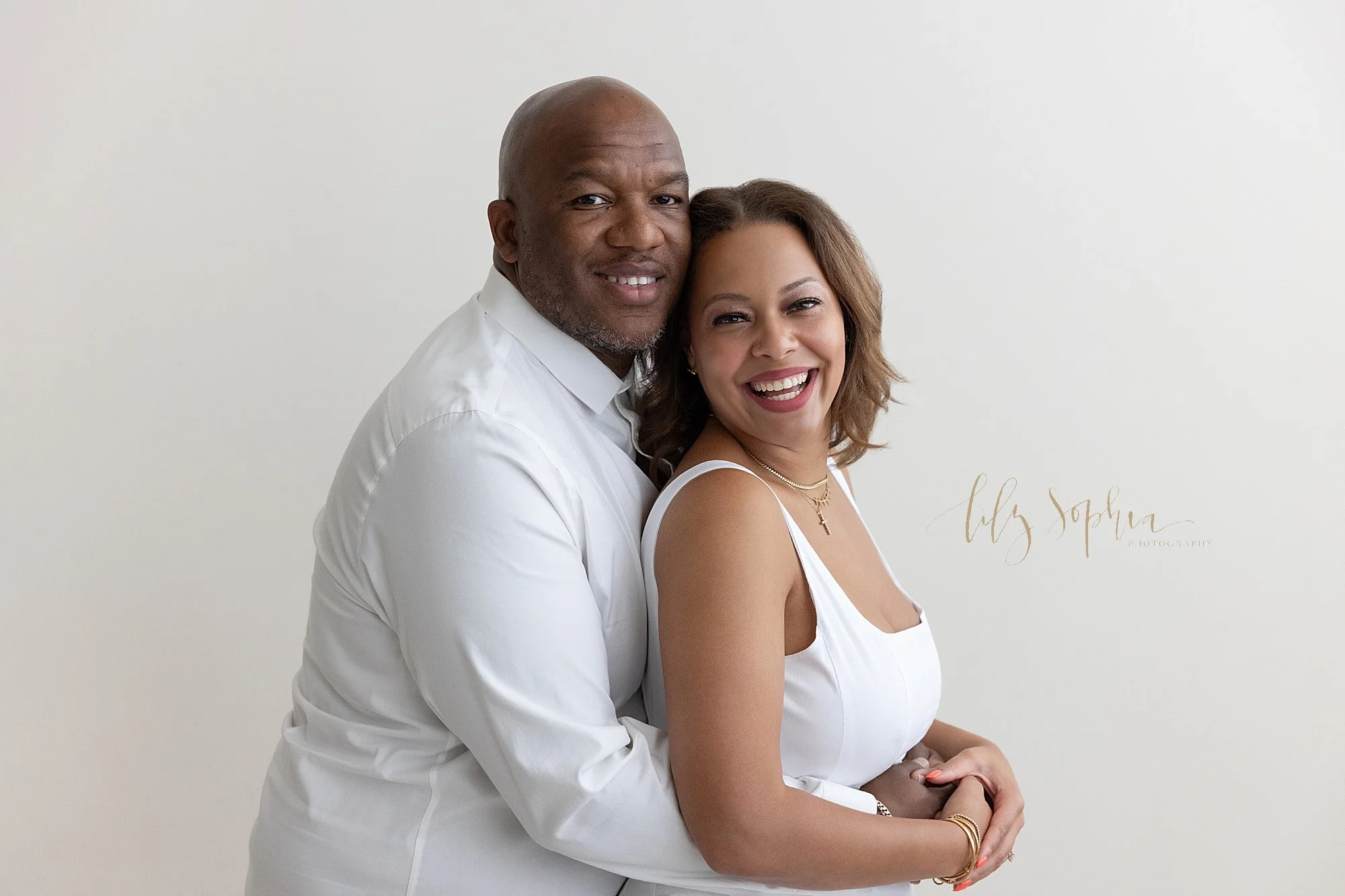  A family photo session with a black husband and wife as he wraps his arms around her waist while she stands with her back against his chest taken in a natural light photography studio near Oakhurst in Atlanta, Georgia 