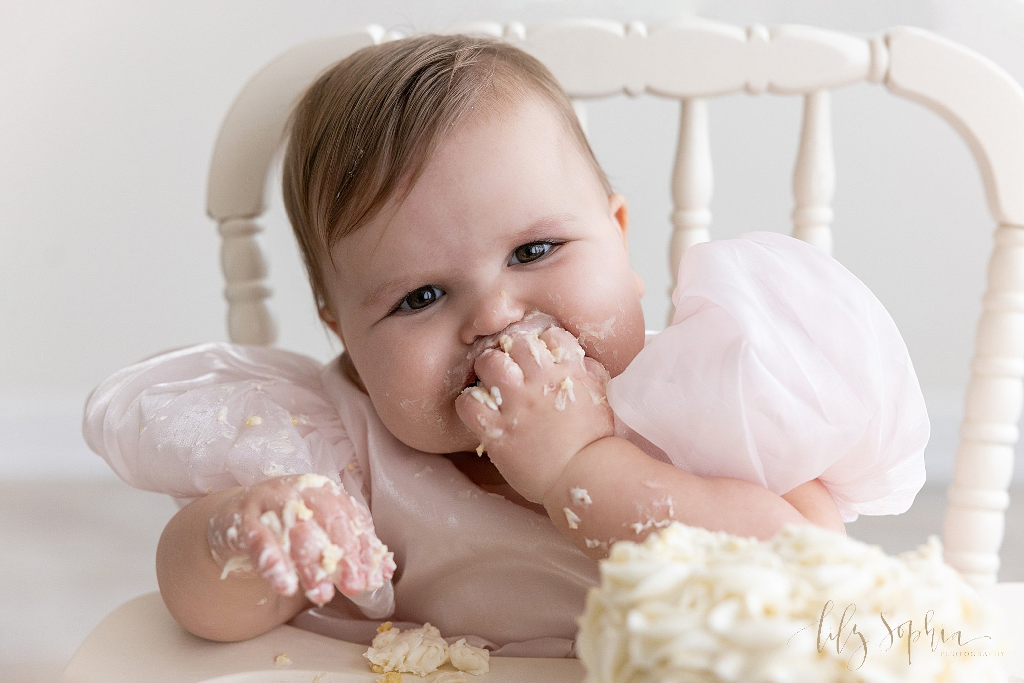  One year old baby girl celebrates her first birthday with a smash cake photo session as she sits in an antique high chair and shoves cake into her mouth in a natural light photography studio located near Cumming in Atlanta, Georgia. 