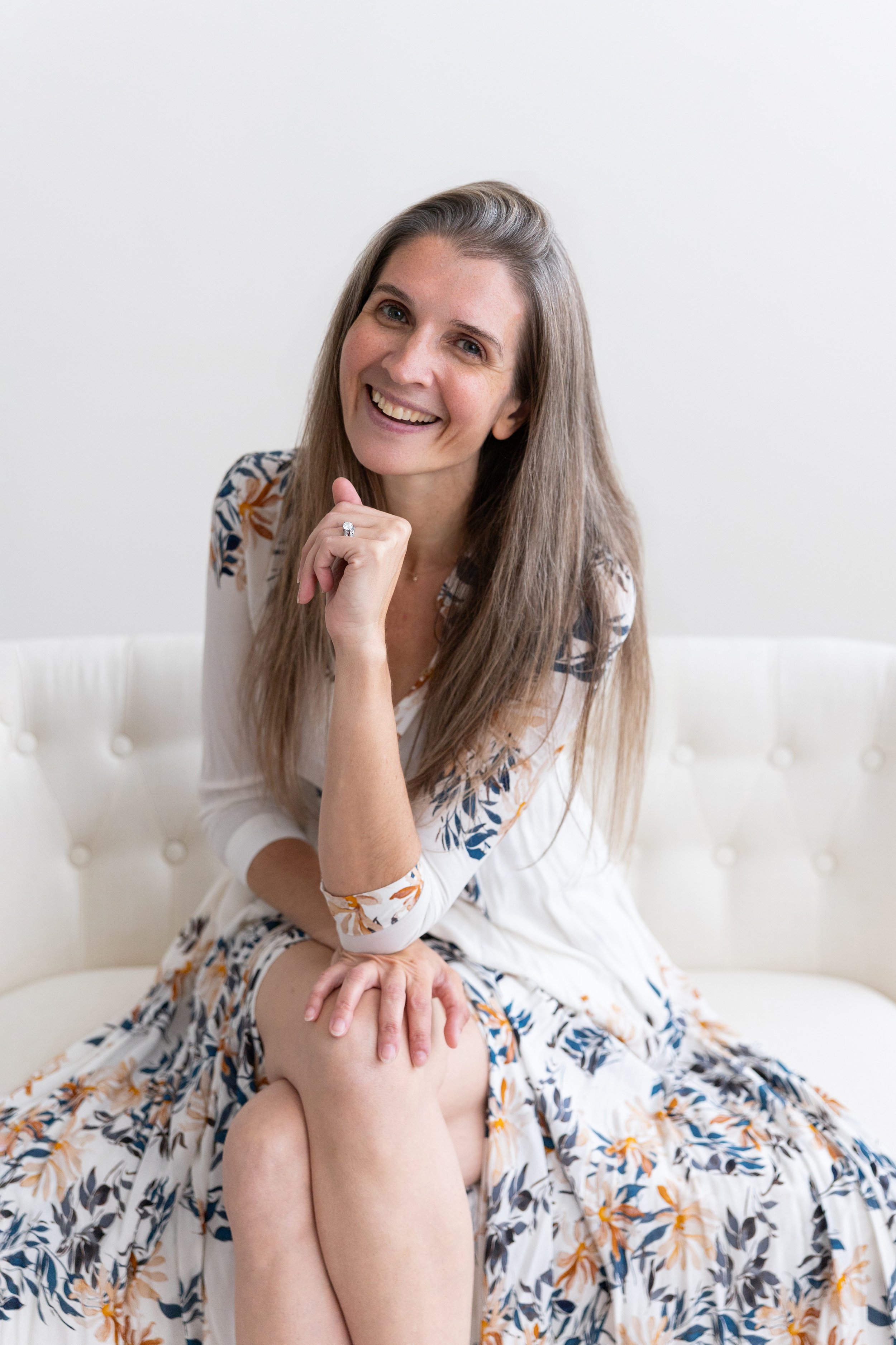 Lauren is the maternity and newborn photographer behind Lily Sophia Photography in Atlanta, Georgia. She is wearing a floral dress while smiling and sitting on a cream couch.