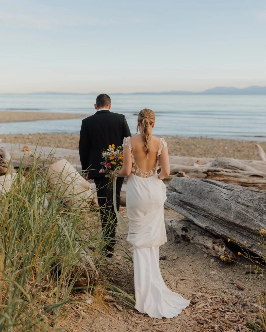 A bride and groom walking on a beach, with the bride holding a colourful bouquet, facing the water during sunset in Gibsons, on the Sunshine Coast.