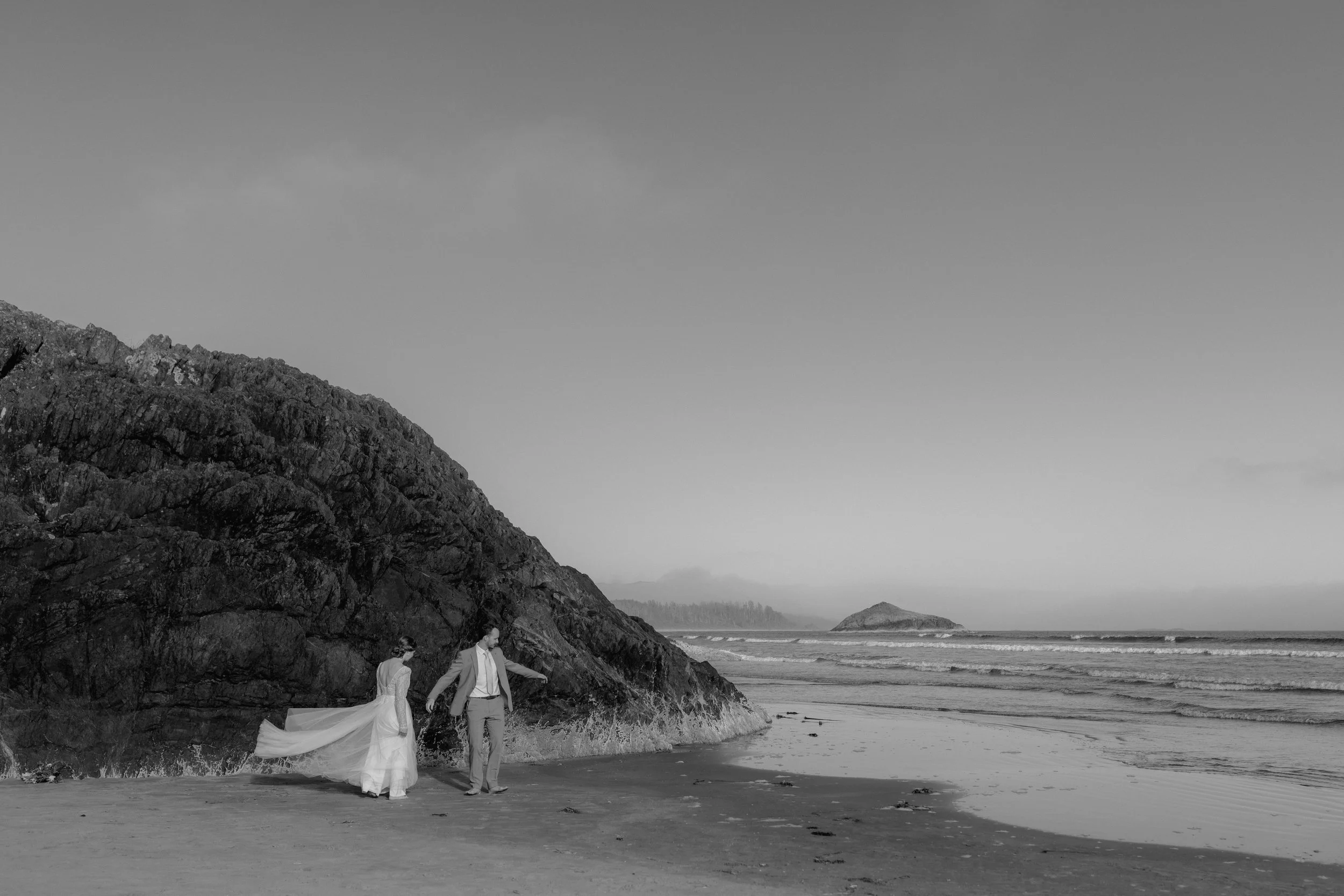 A black and white photo of a couple on a beach, walking along the shoreline near a large rocky hill, with ocean waves in the background.
