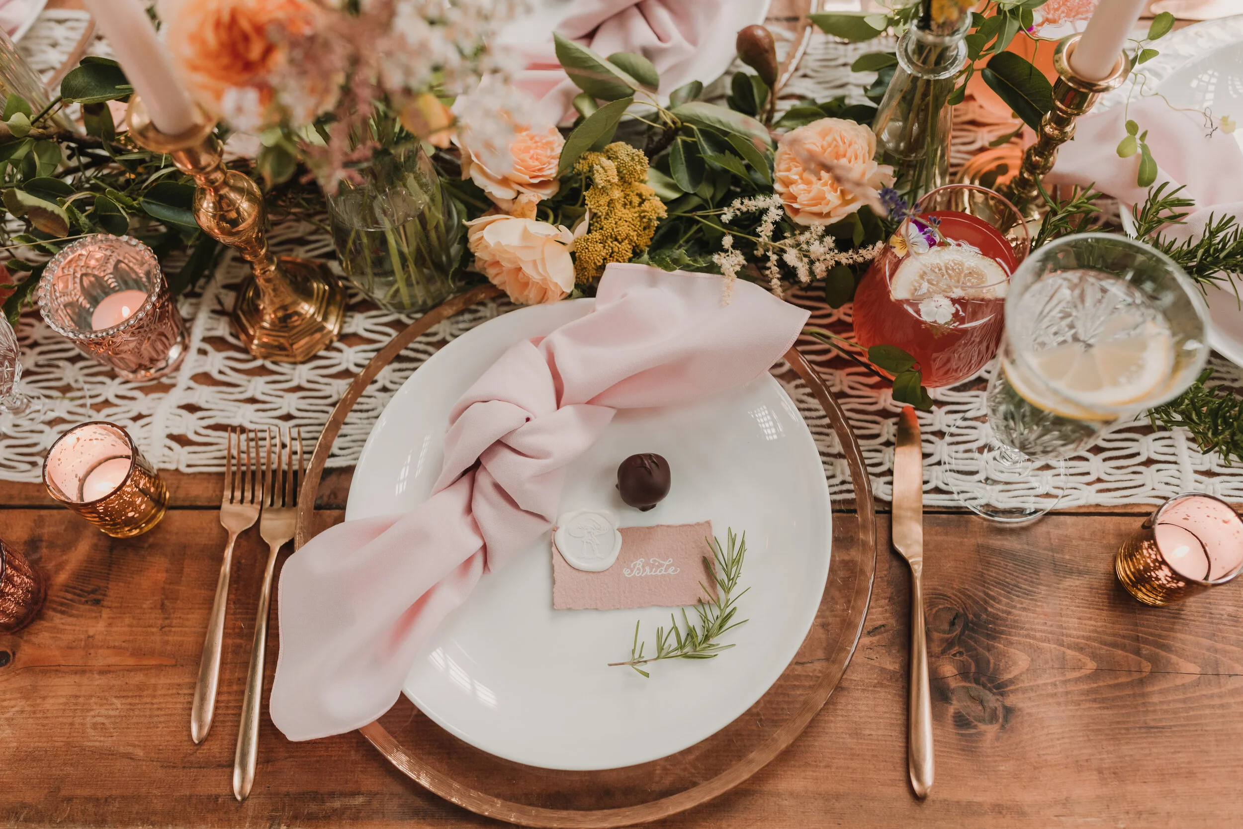 Elegant wedding table setting with a white plate, pink cloth napkin, chocolate, and a small pink 'bride' place card. Surrounding the plate are pink and gold candle holders, flowers, and greenery on a wooden table with a lace runner.