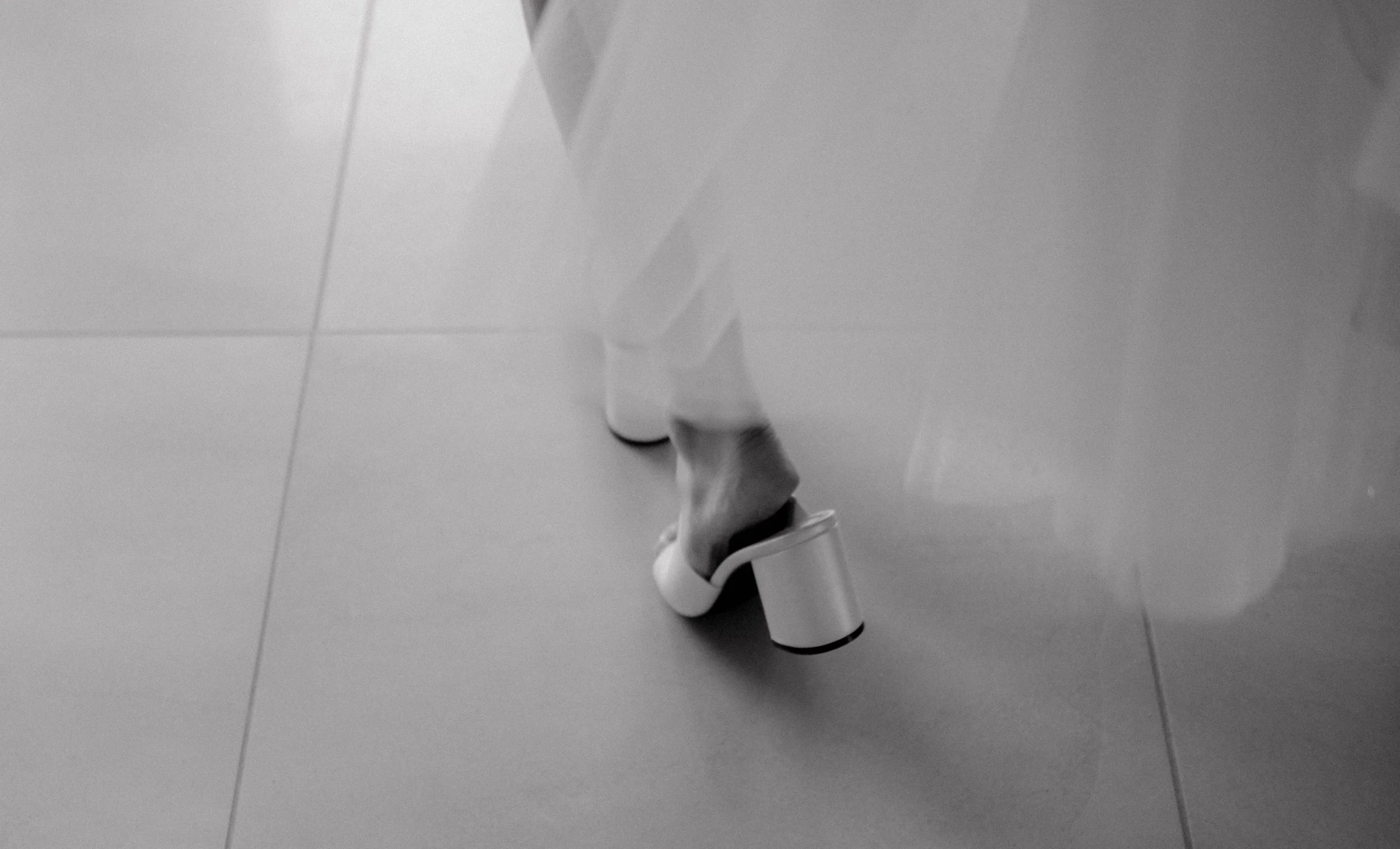 Close-up of a person's foot wearing a white shoe with a thick heel, standing on a tiled floor next to a reflective surface.