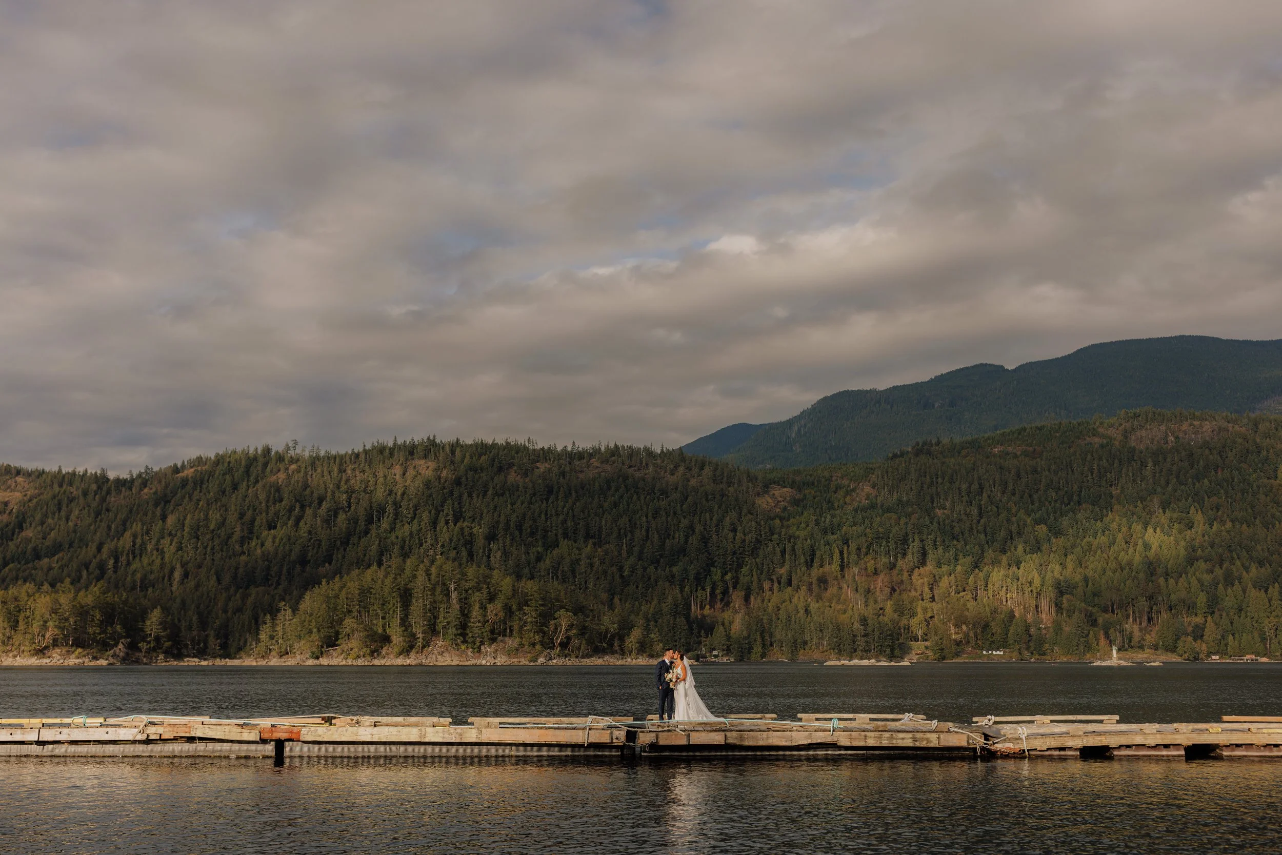 A bride and groom standing on a wooden dock by a lake, with forested hills and mountains in the background under a partly cloudy sky.