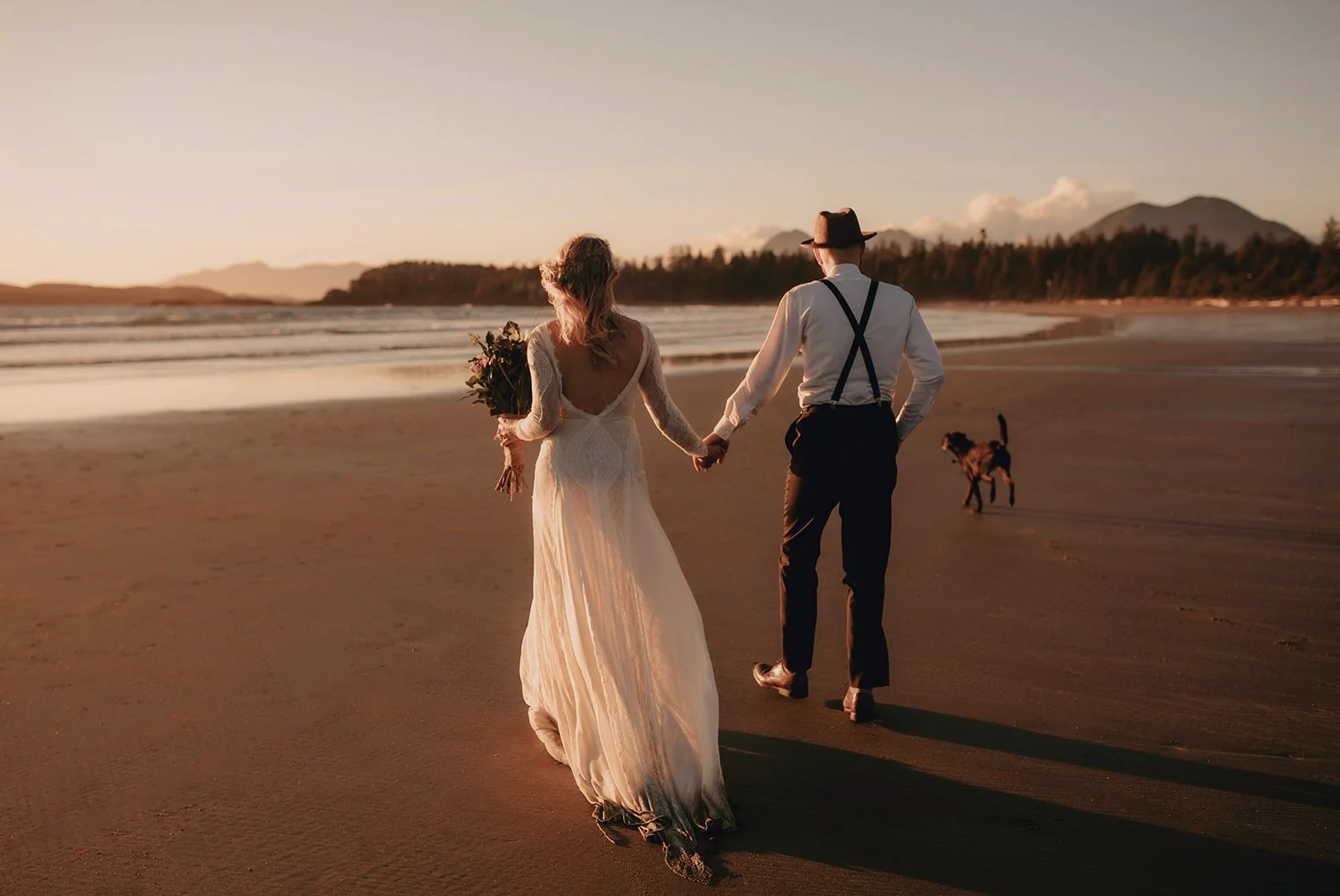 A couple in wedding attire walking hand in hand on a sandy beach at sunset, with their dog running beside them and mountains in the background on Chesterman Beach, Tofino.