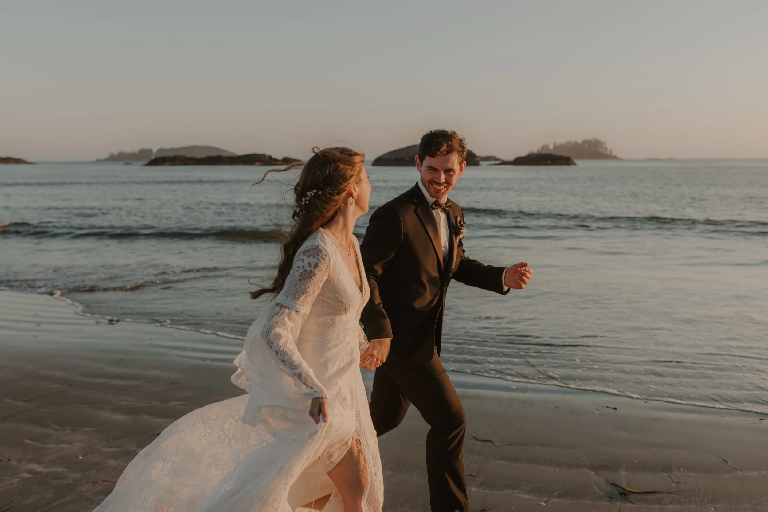 A newlywed couple running hand in hand along the beach at sunset, with the ocean and small islands in the background at Mackenzie Beach in Tofino, BC