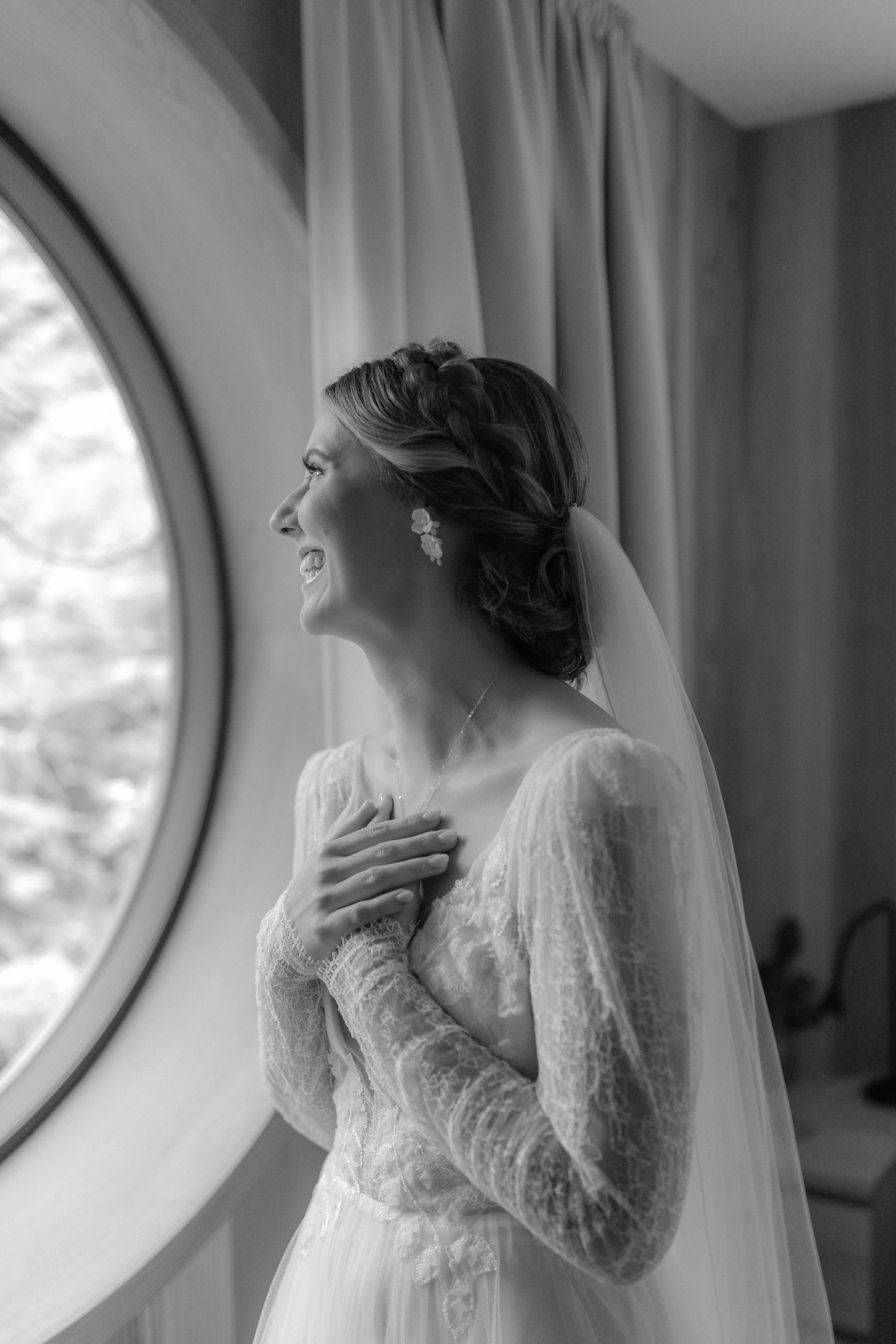 A bride in a Lacey wedding dress, and earrings looks out a large round window, smiling with her hand on her chest in a black and white photo in her getting ready suite in Tofino, BC