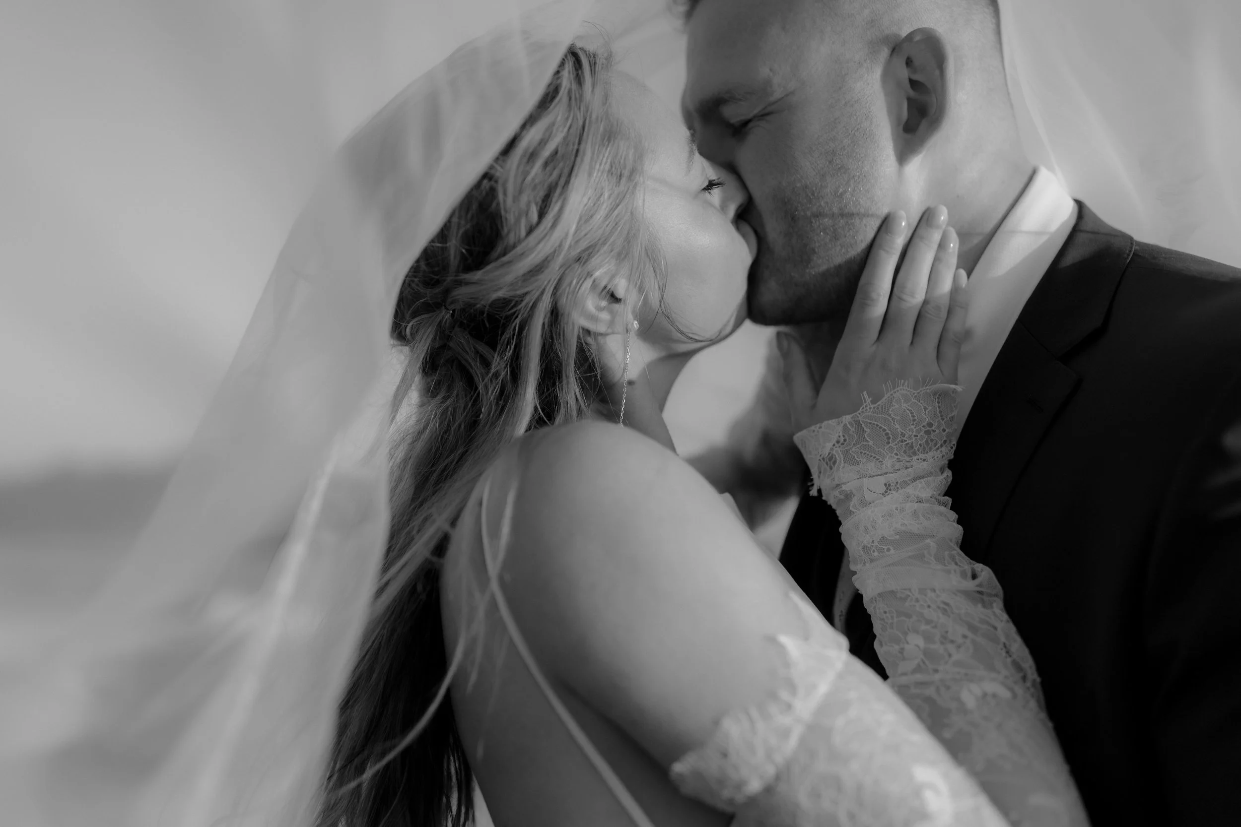 A bride and groom share a kiss on their wedding day, with the bride wearing a veil and lace dress, and the groom in a suit.