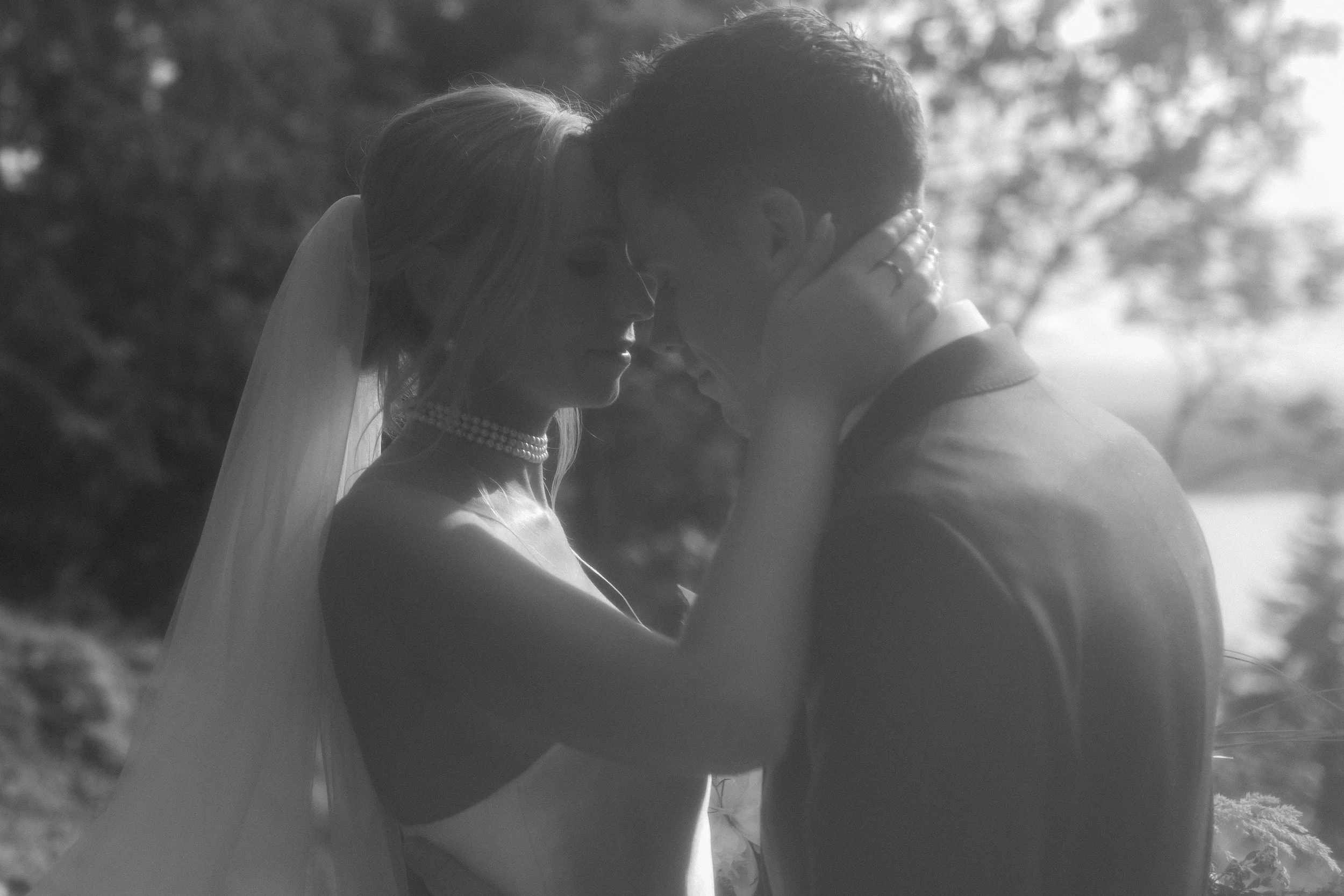 A black and white photo of a bride and groom with their foreheads and noses touching, embracing outdoors with trees and sky in the background.