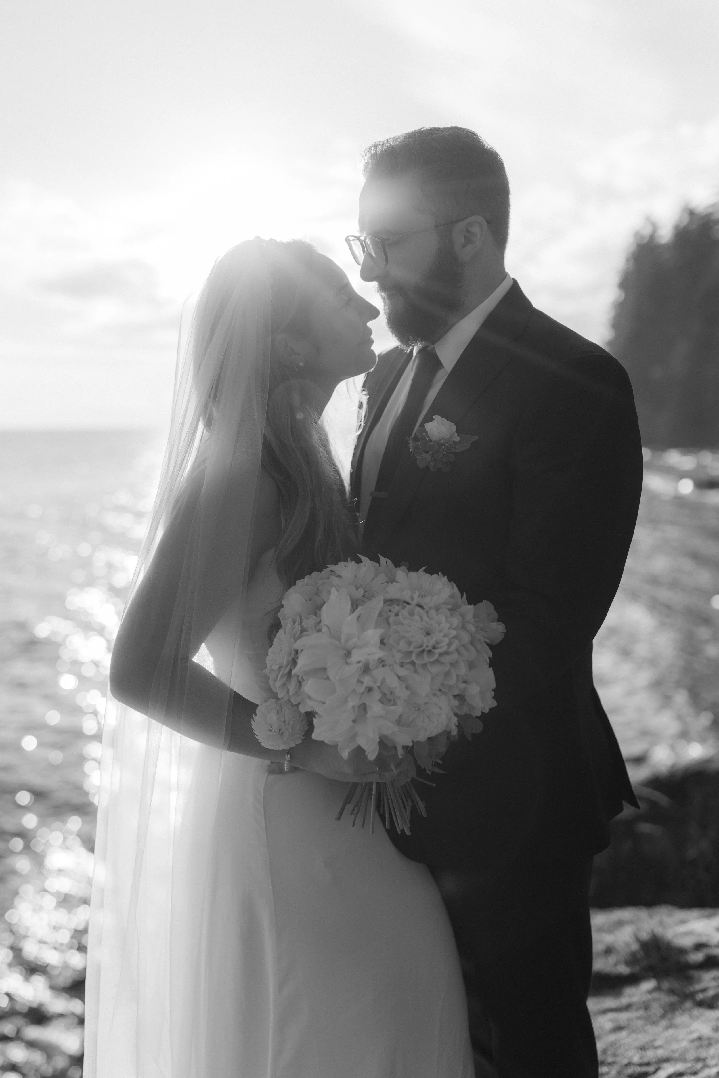 A bride and groom stand close together by the water at sunset, smiling and gazing at each other, with the bride holding a bouquet of flowers.