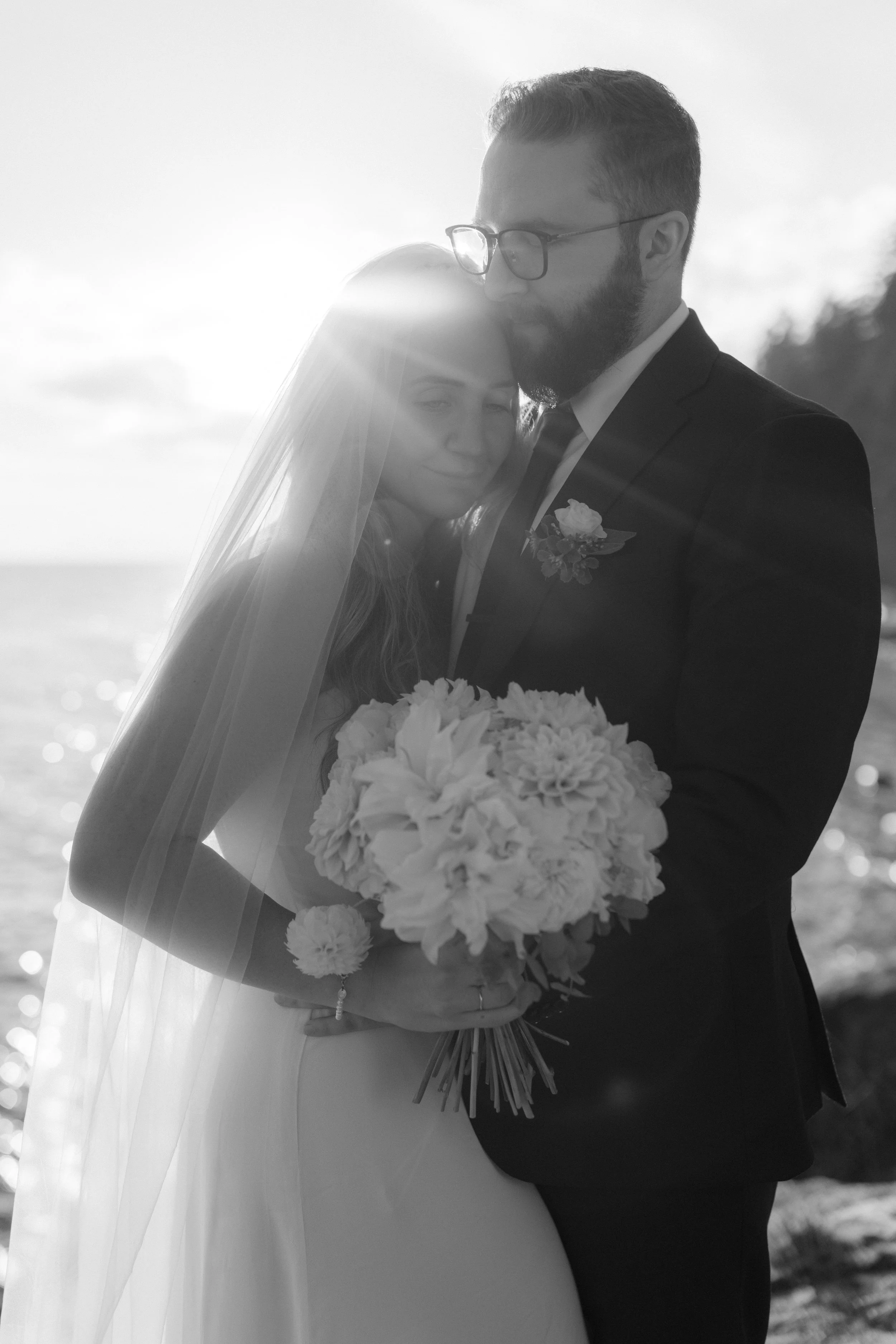Black and white photo of a wedding couple embracing by the water, with the sunlight creating a halo effect around them. The bride is holding a bouquet of flowers, and both are dressed in formal wedding attire on the Sunshine Coast.