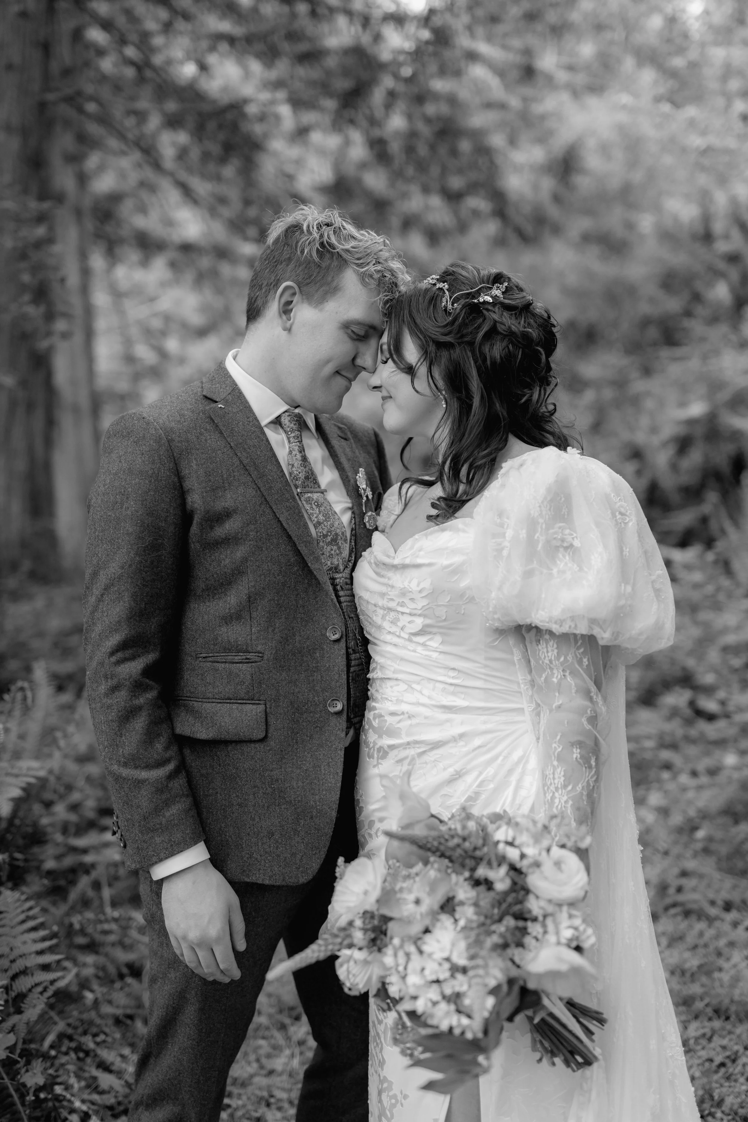 A black and white photo of a bride and groom standing outdoors in a forest setting, touching foreheads and smiling, with the bride holding a bouquet of flowers.