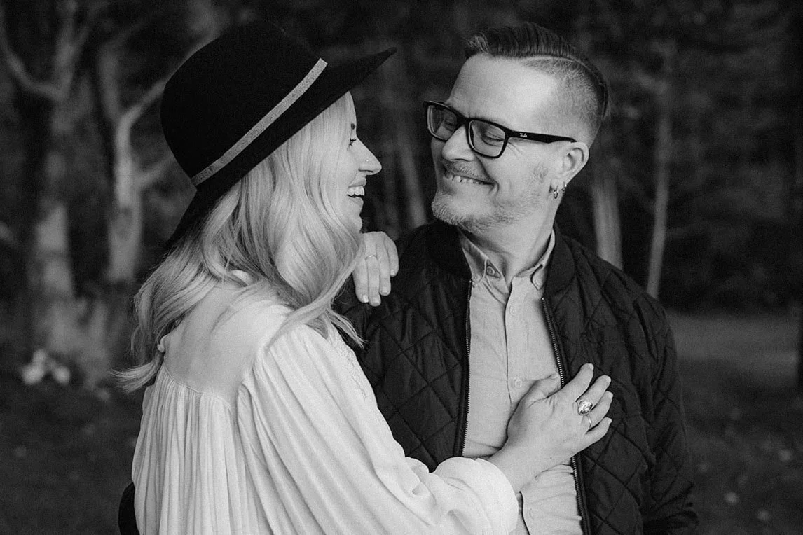 A couple smiling and looking at each other outdoors, with the woman wearing a wide-brimmed hat and the man wearing glasses.