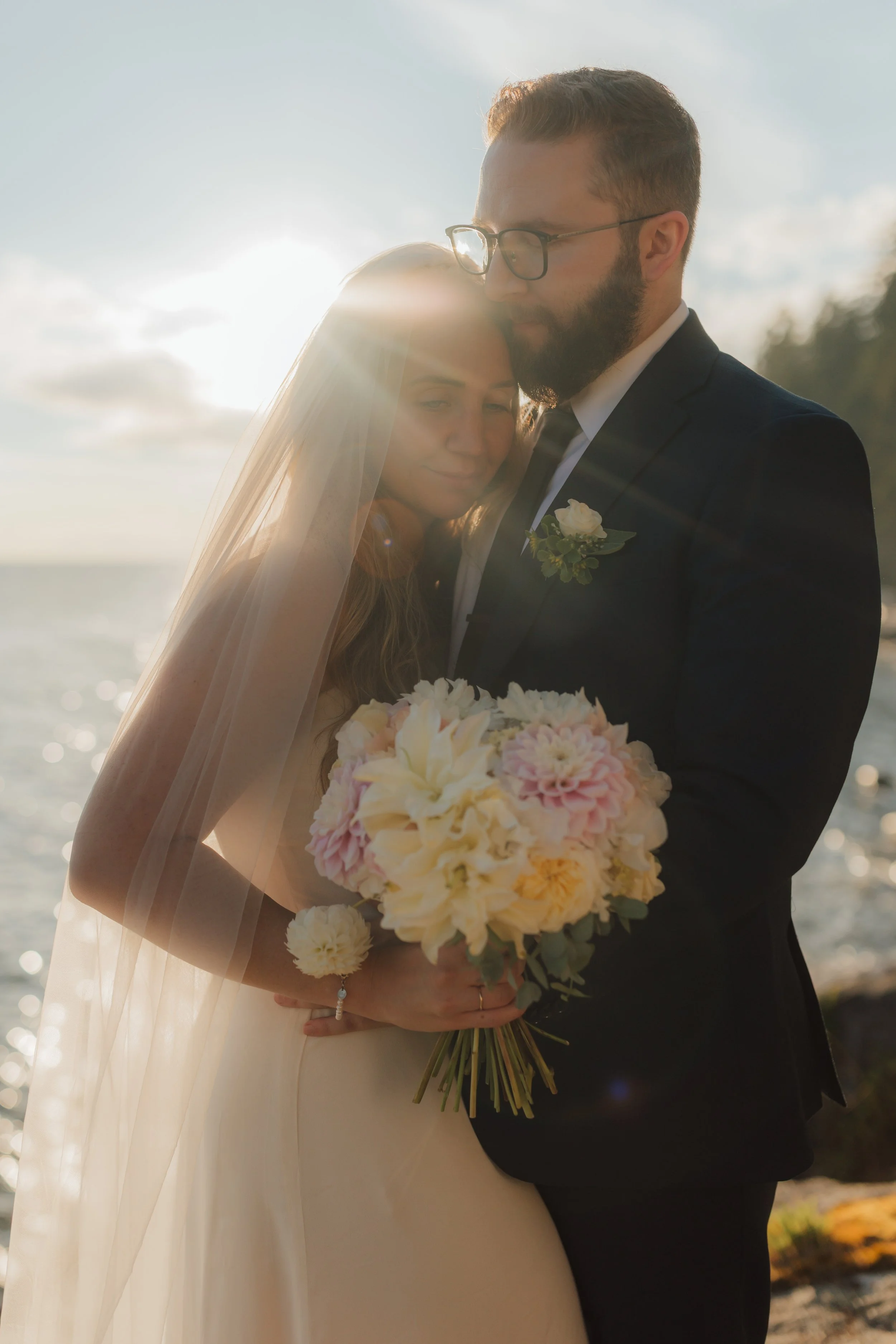 A bride and groom standing close together outdoors at sunset, holding a bouquet of flowers, with the sun behind them creating a backlit effect.