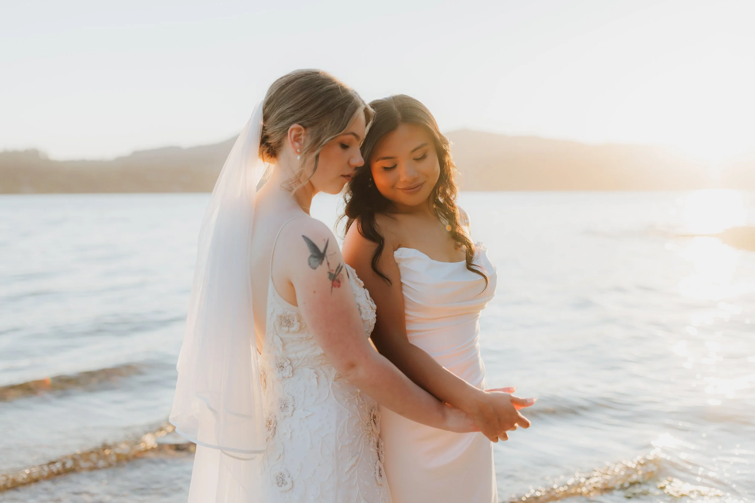 Two women in wedding dresses standing at the water's edge during sunset, holding hands and looking down, with calm water and a distant shoreline in the background on the Sunshine Coast.